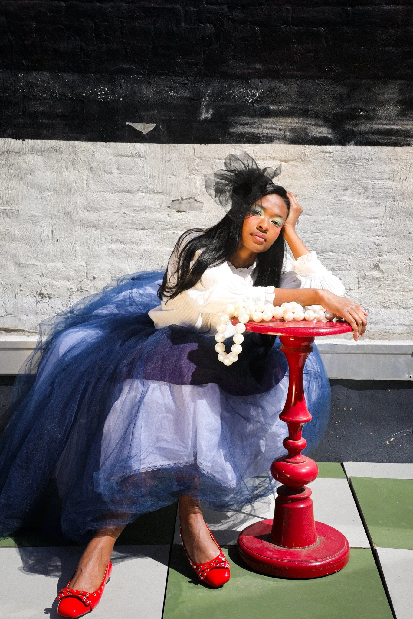 Woman dressed in a colorful, flowing dress with a large black mesh fascinator on her head, posing against a black and white painted brick wall, leaning on a red side table with a string of large white pearls, wearing red high heels with silver embellishments.