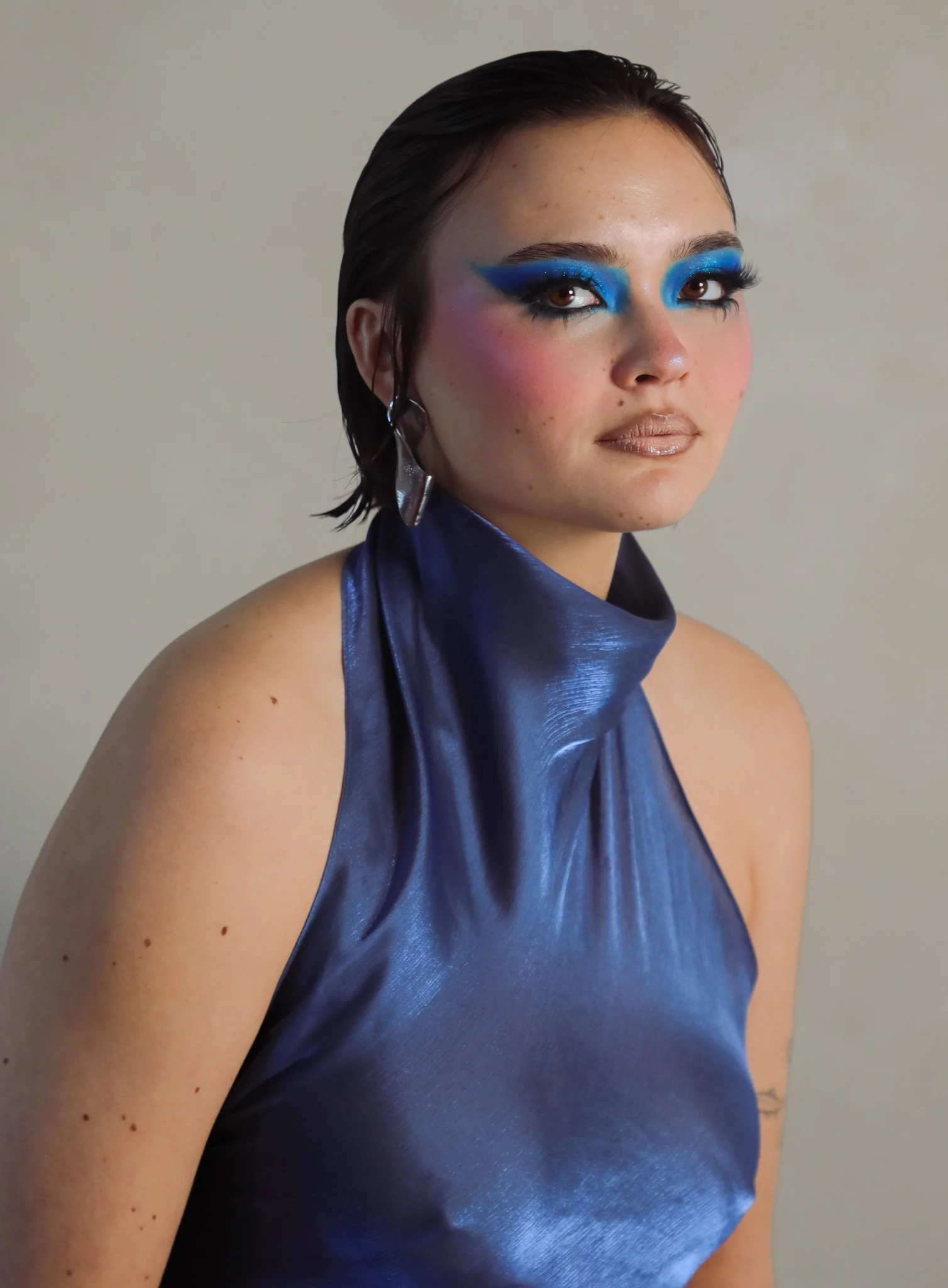A woman with dark wet hair, wearing blue eyeshadow, dramatic eyeliner, glitter, and a shiny blue satin halter top.
