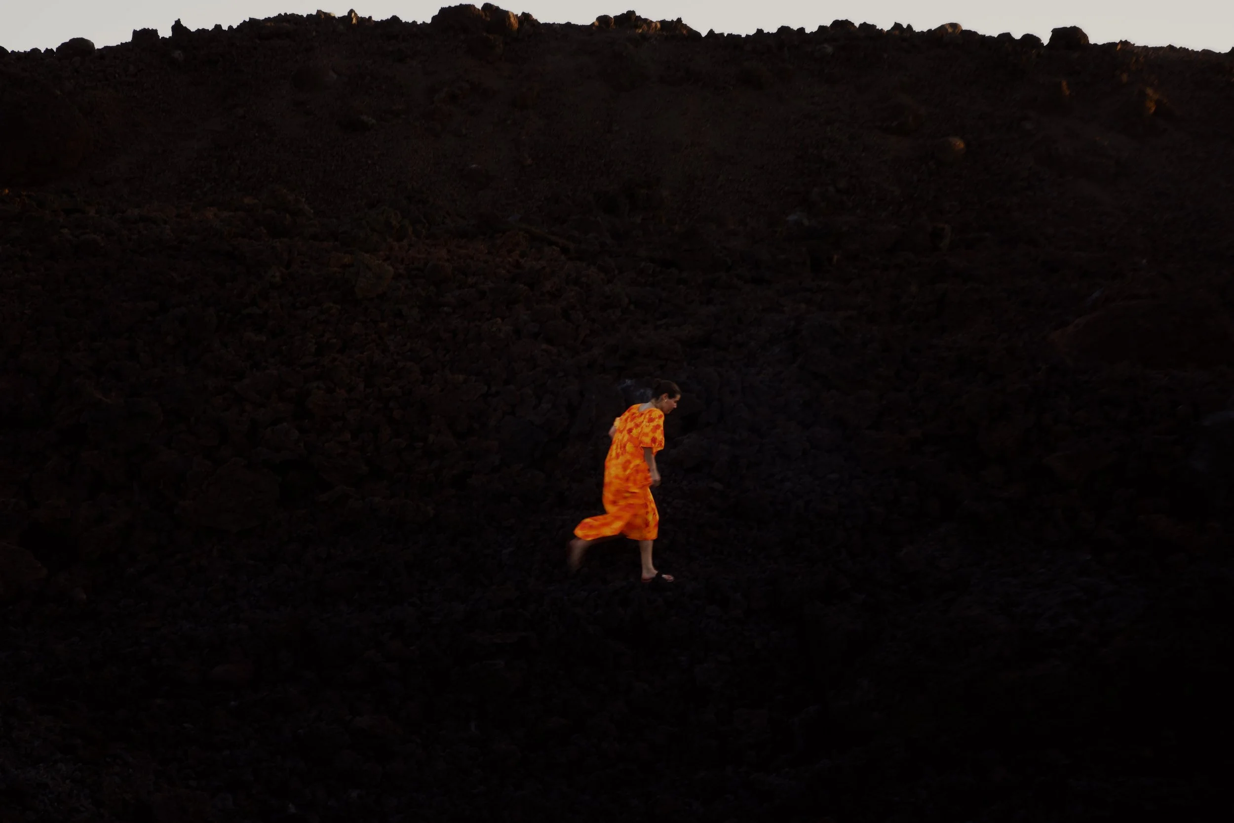 Person wearing a bright orange dress walking barefoot on dark dirt or volcanic rock terrain with a large hill or mountain in the background during dusk or dawn.
