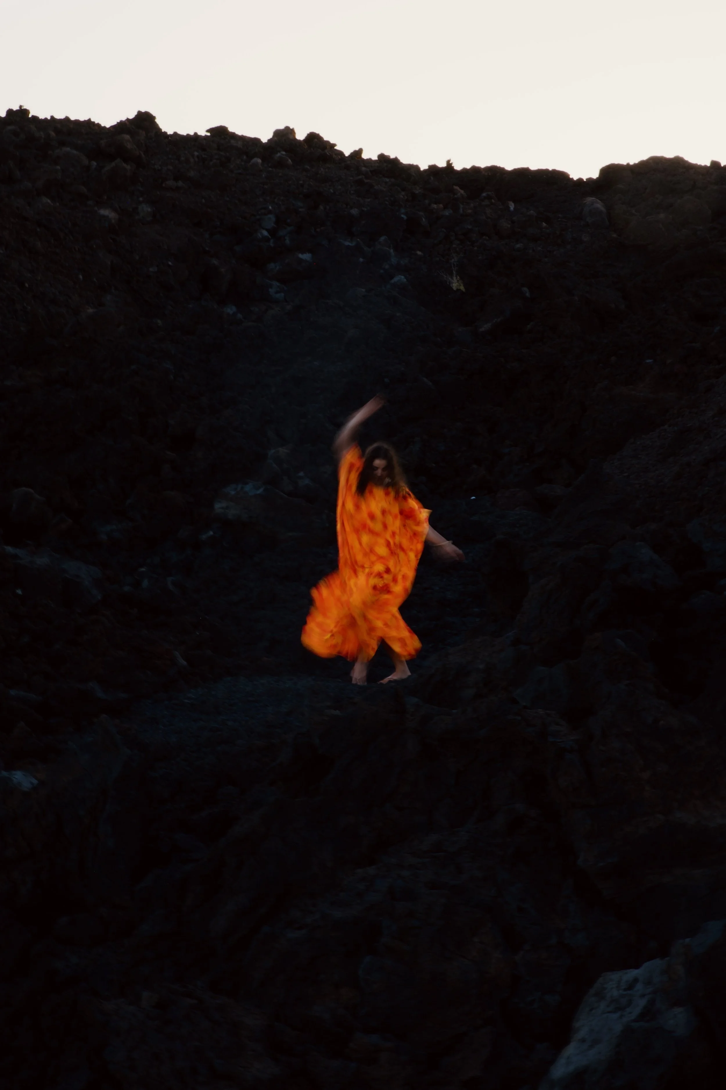 A person in an orange dress dancing on black volcanic rocks with a bright sky in the background.