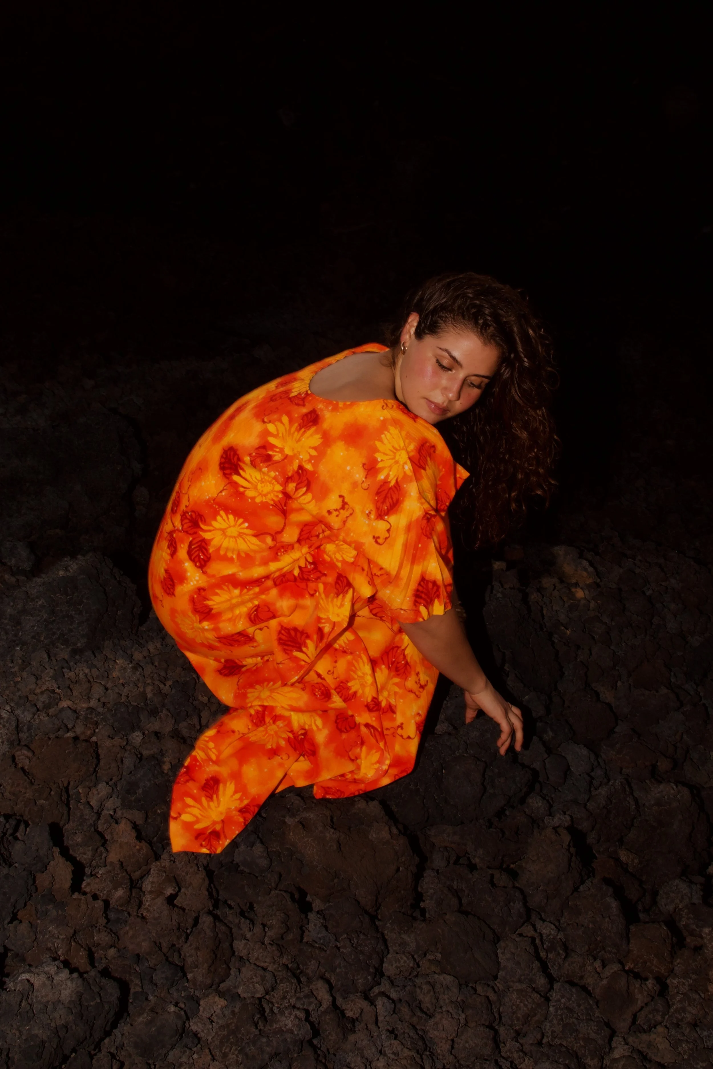 A woman with curly hair, dressed in an orange floral dress, is sitting on dark, volcanic rock at night, looking down with her eyes closed.