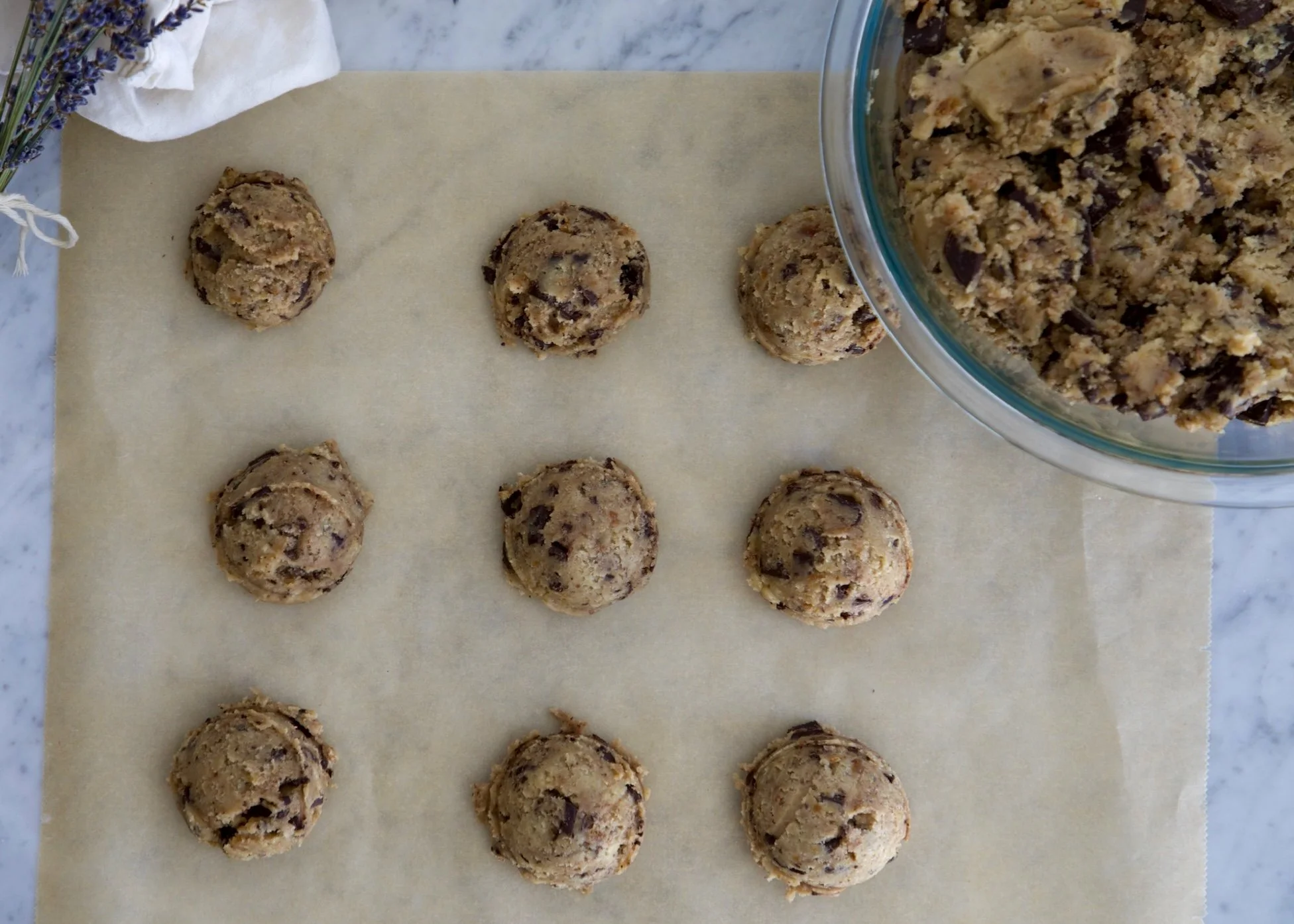 Freshly baked cookie dough balls with chocolate chips on parchment paper and a large bowl of cookie dough to the side.