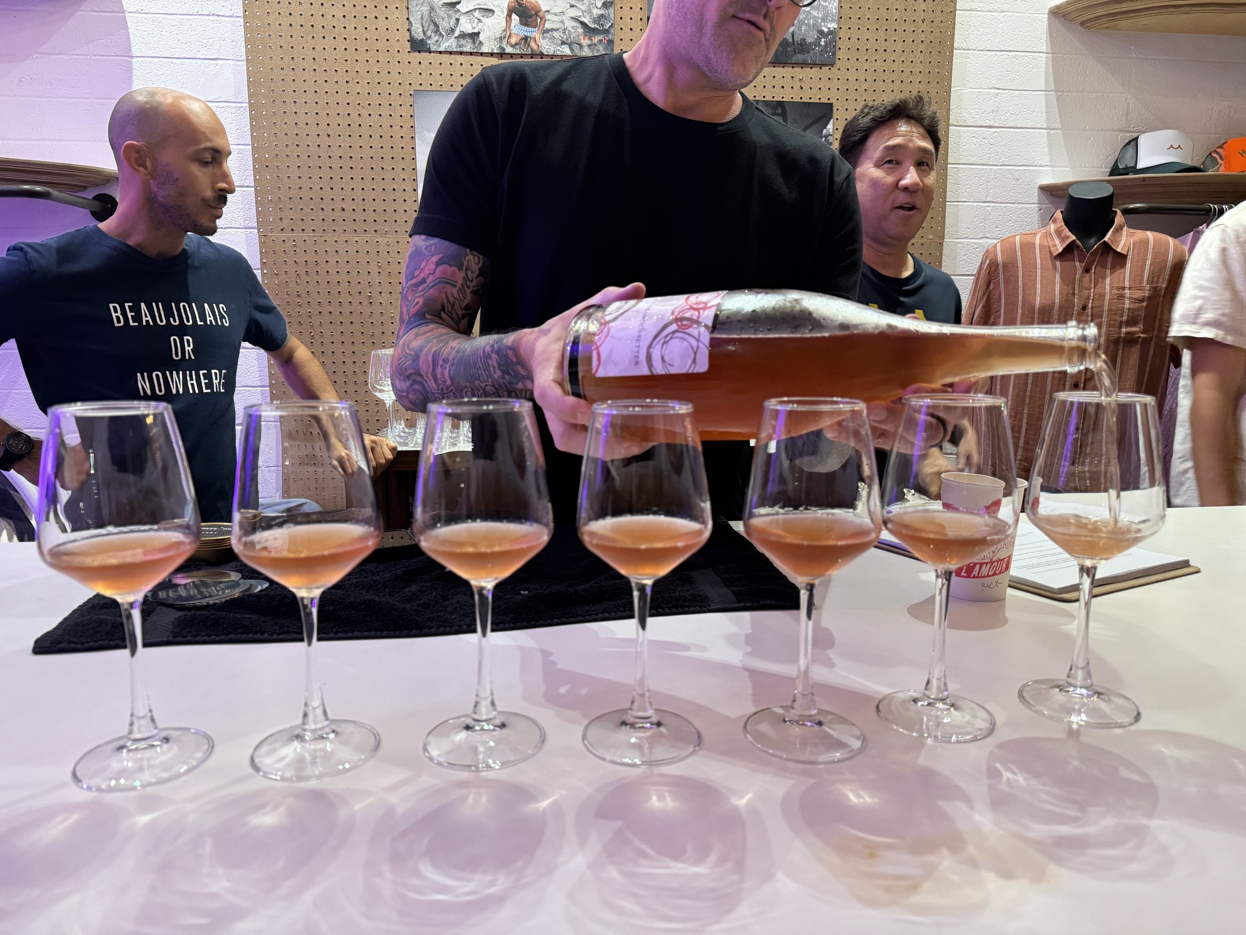 A man pours rosé wine into glasses on a white table, surrounded by three other men, with a pegboard and photos on the wall in the background.