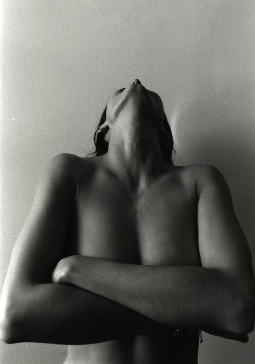 Black and white photo of a topless woman with arms crossed, head tilted back, and eyes closed against a plain wall.