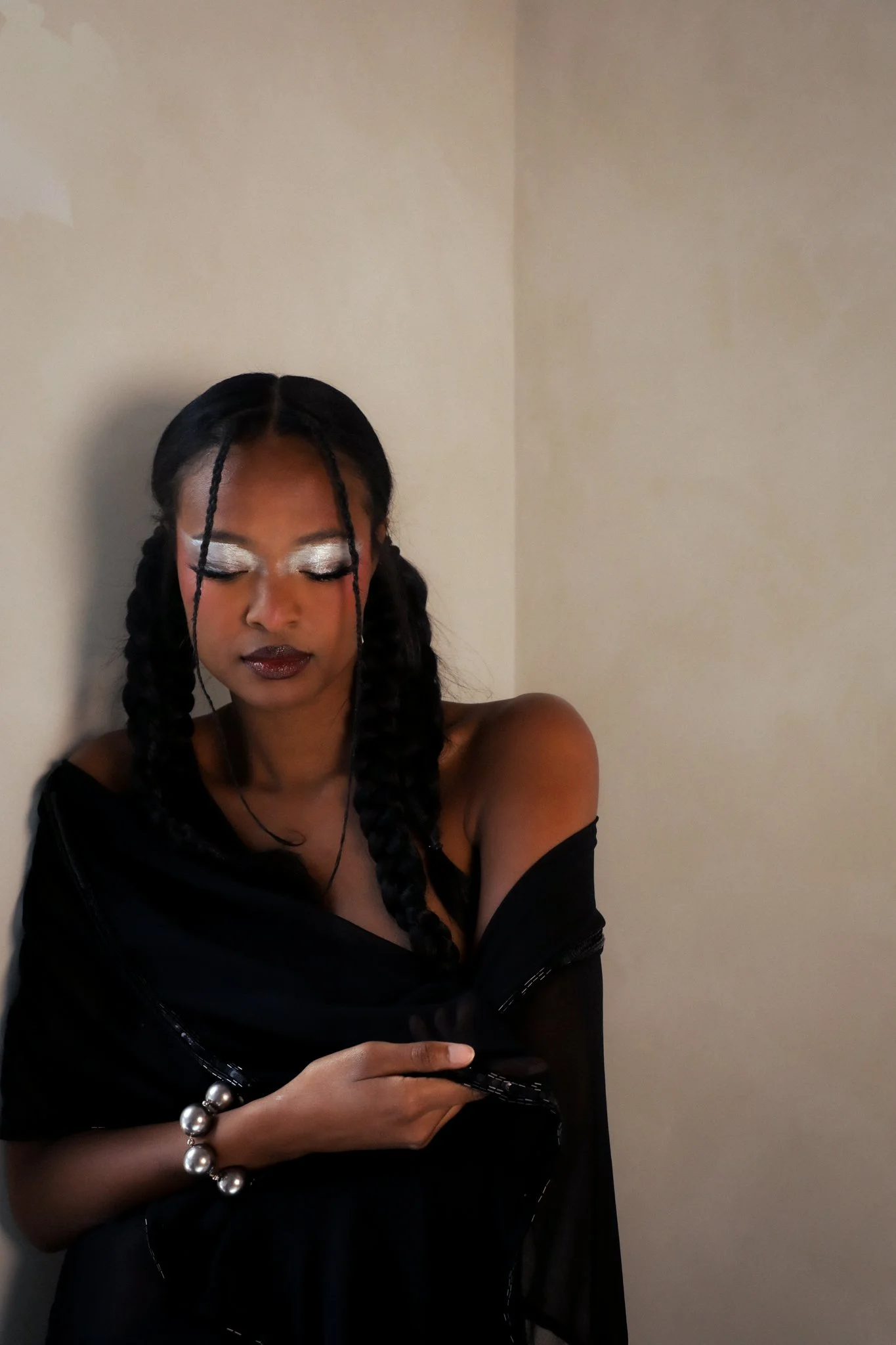A woman with long braided hair and silver eye makeup taking a selfie against a beige wall. She is wearing a black outfit and silver jewelry, with a contemplative or relaxed expression.
