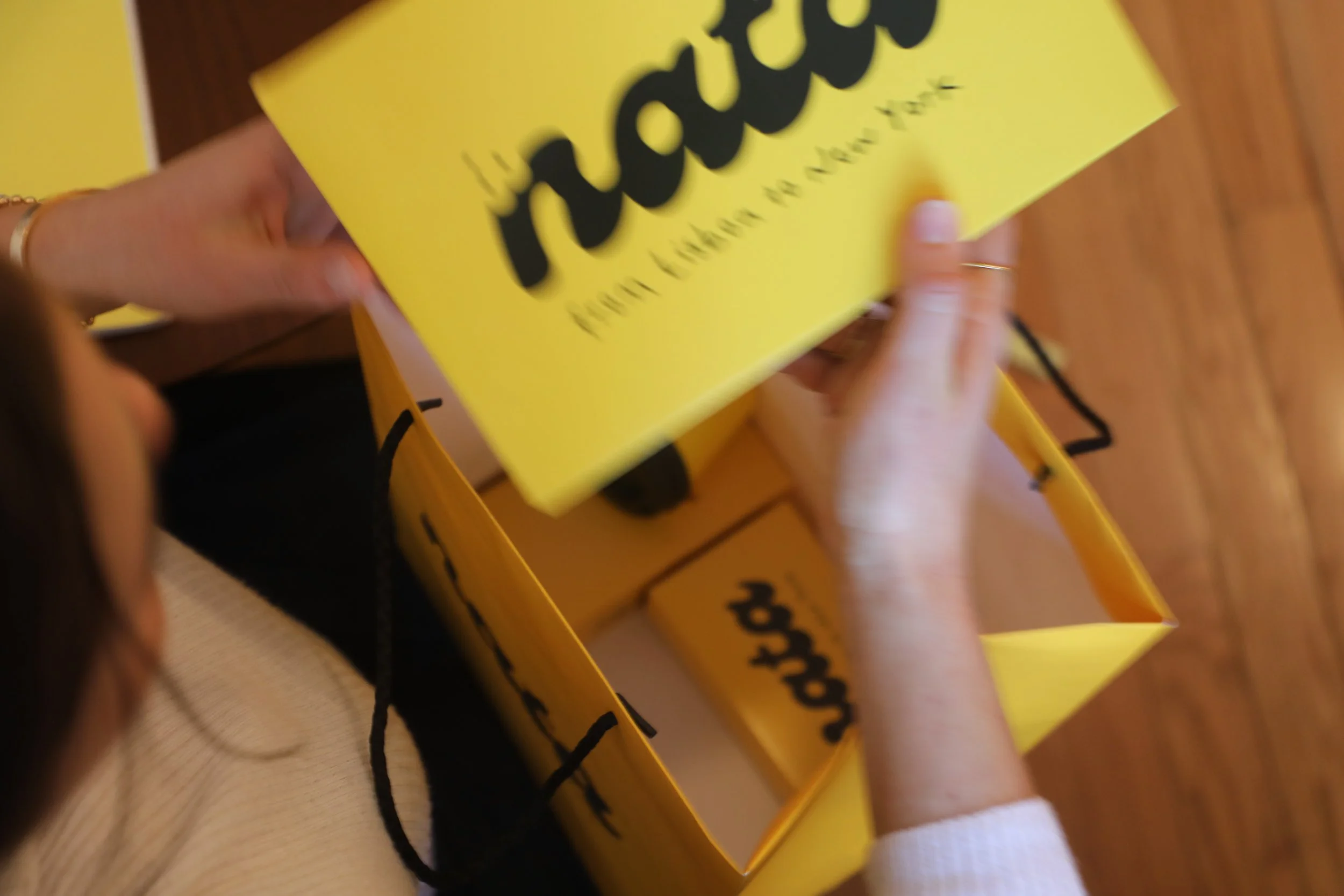 Close-up of a person's hand holding a small yellow box with black text and a large yellow sign with black text. The background is a wooden surface.