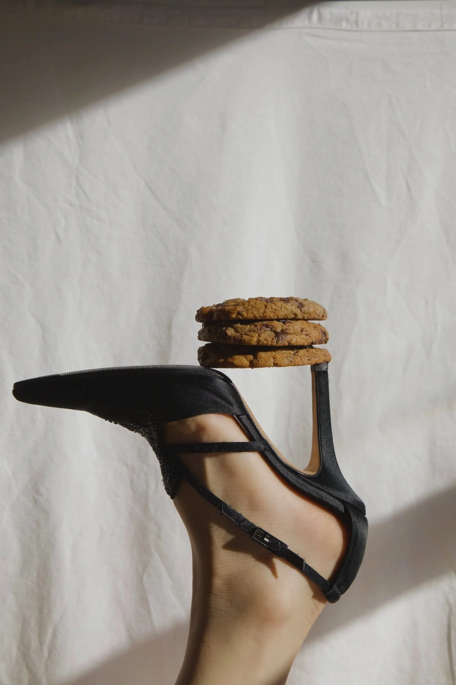 A hand wearing a black strappy high heel shoe holding a stack of three chocolate chip cookies against a plain white background.