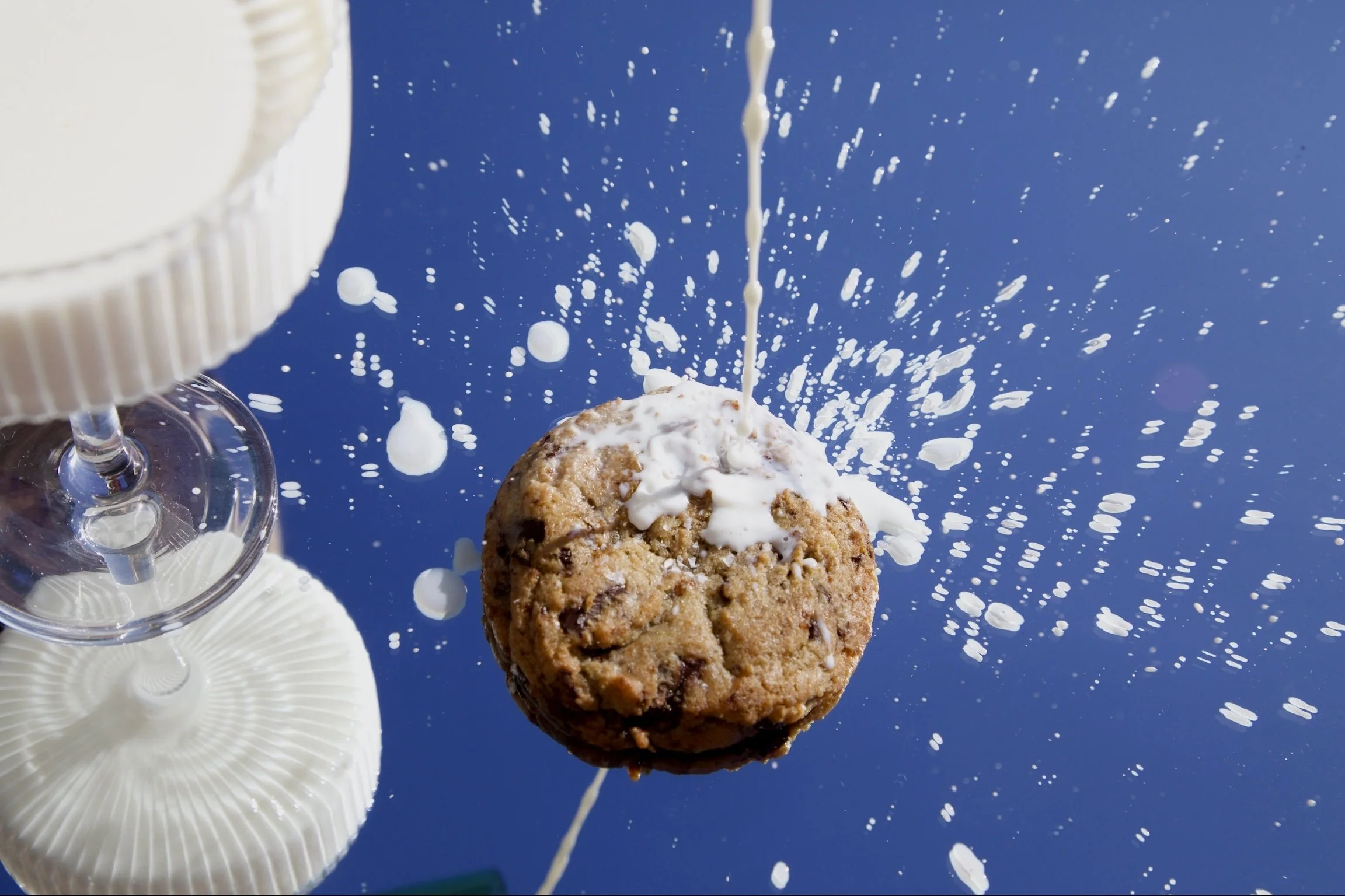 A chocolate chip cookie with white icing being drizzled over it, with a few cookies and a white cupcake liner in the foreground, against a bright blue background with falling white sprinkles.