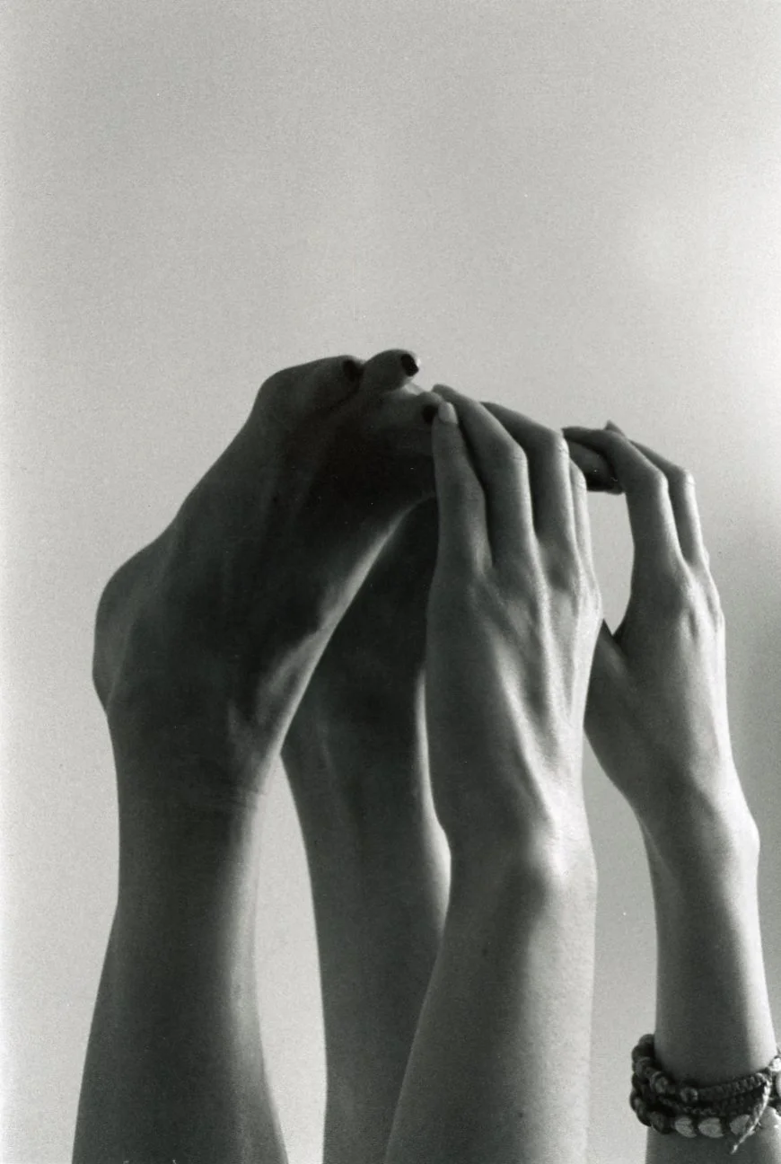 Black and white photo of hands reaching up and holding each other, with one hand wearing a beaded bracelet.