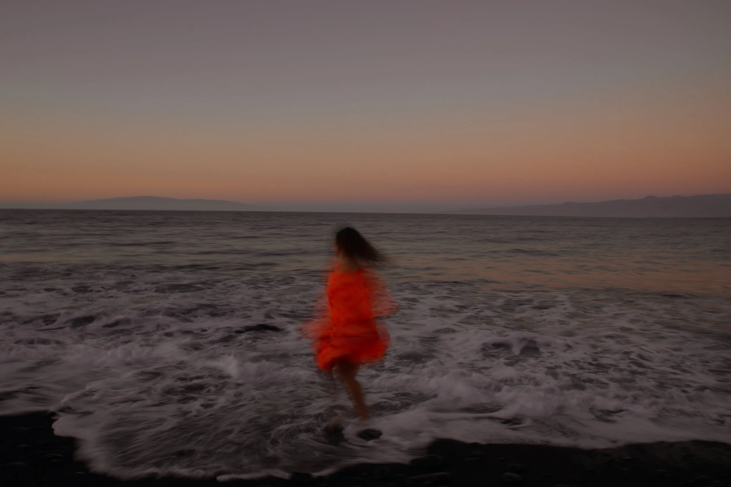 A person running in the ocean at sunset, wearing a red outfit with a blurred background of the water and distant land.