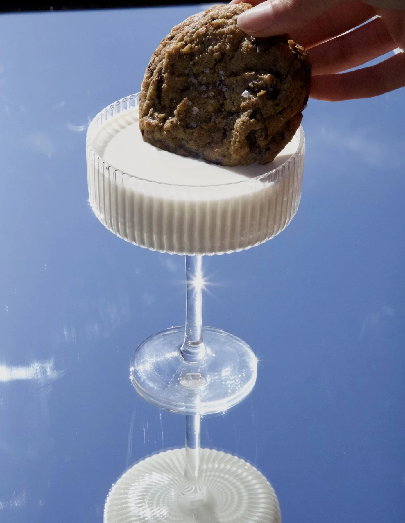 A hand holding a cookie over a clear cocktail glass filled with a white beverage, with reflections on the glass and a blue sky background.
