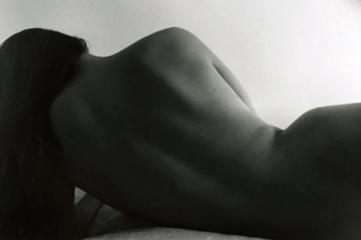 Black and white photo of a woman lying sideways on a flat surface, with her back and shoulder visible and her head turned away.