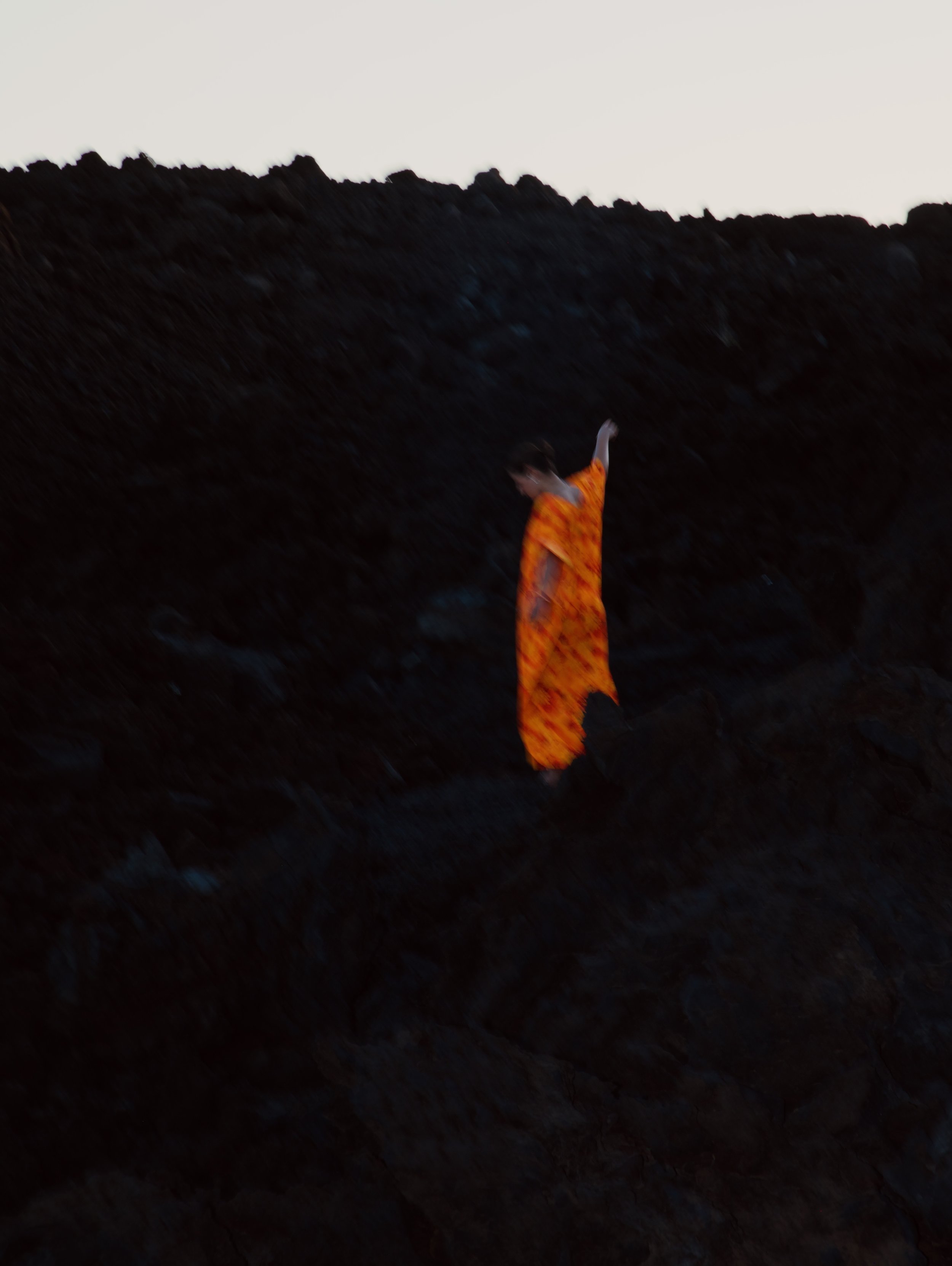 A person in an orange patterned dress with long hair, standing outdoors on a dark, rocky ground with a bright sky in the background.