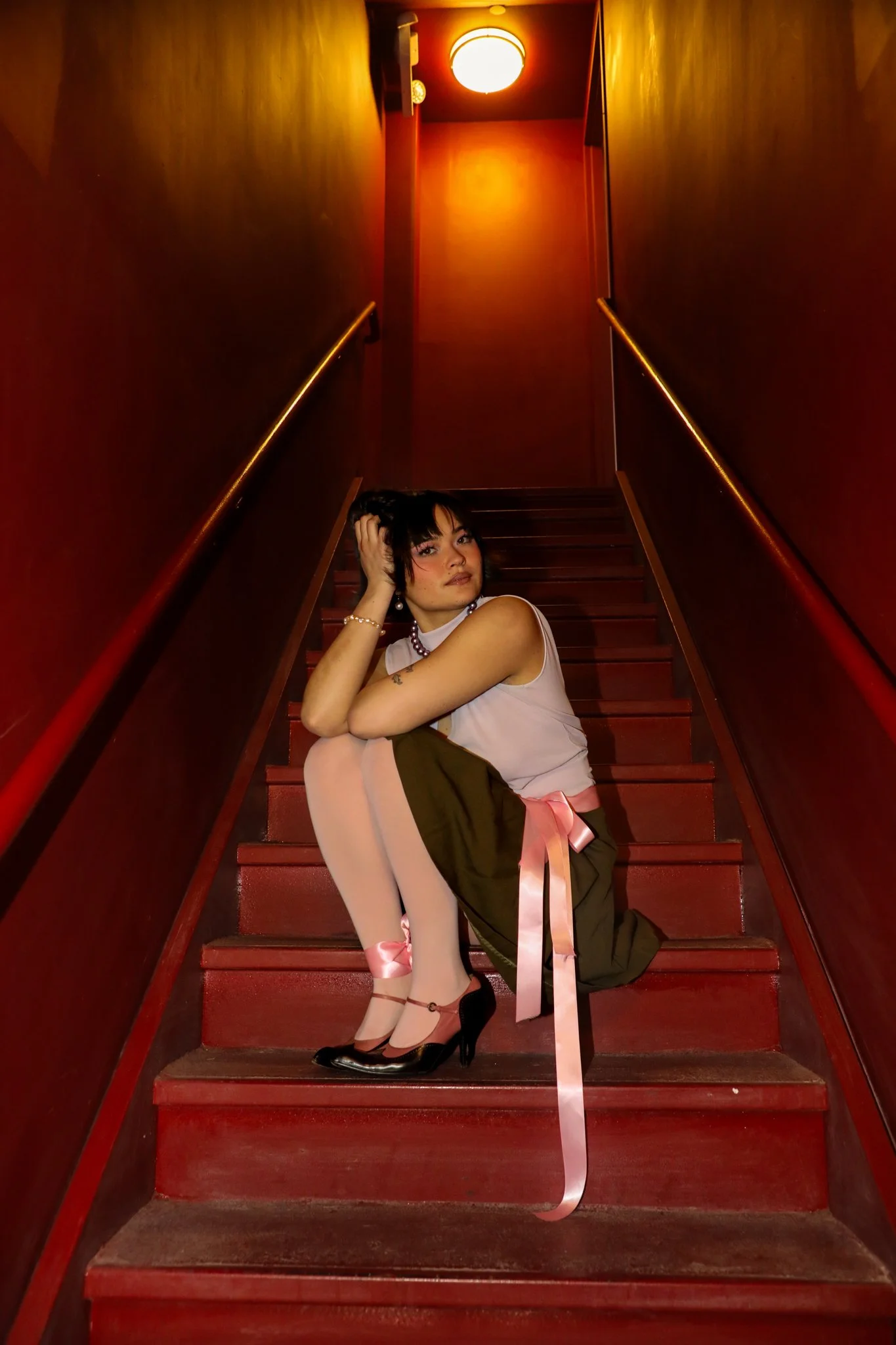 A woman dressed in a vintage-inspired outfit sitting on red stairs with her legs crossed. She is wearing a pink top, a dark skirt with pink satin ribbons, and black high heels with ankle straps. She has short dark hair, makeup, and accessories including a pearl bracelet and earrings. The setting appears to be a dimly lit corridor with red walls and a ceiling light.