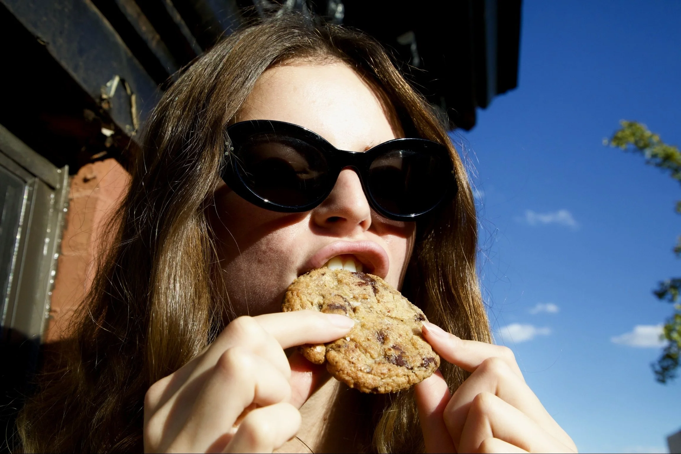 Woman with brown hair wearing black sunglasses eating a large chocolate chip cookie outdoors on a sunny day.