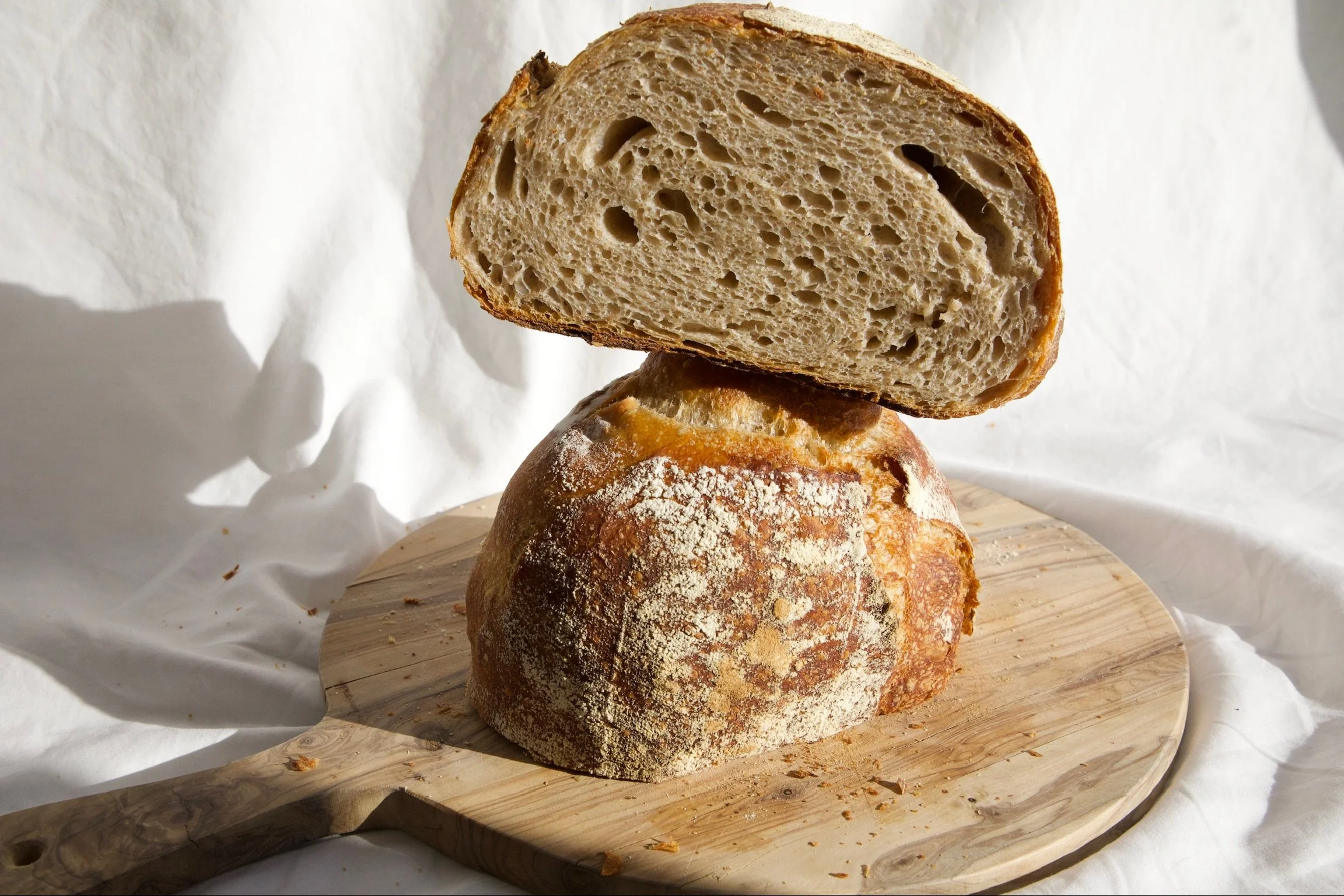A loaf of rustic bread with a soft, airy interior, sitting on a wooden cutting board with a white cloth background.