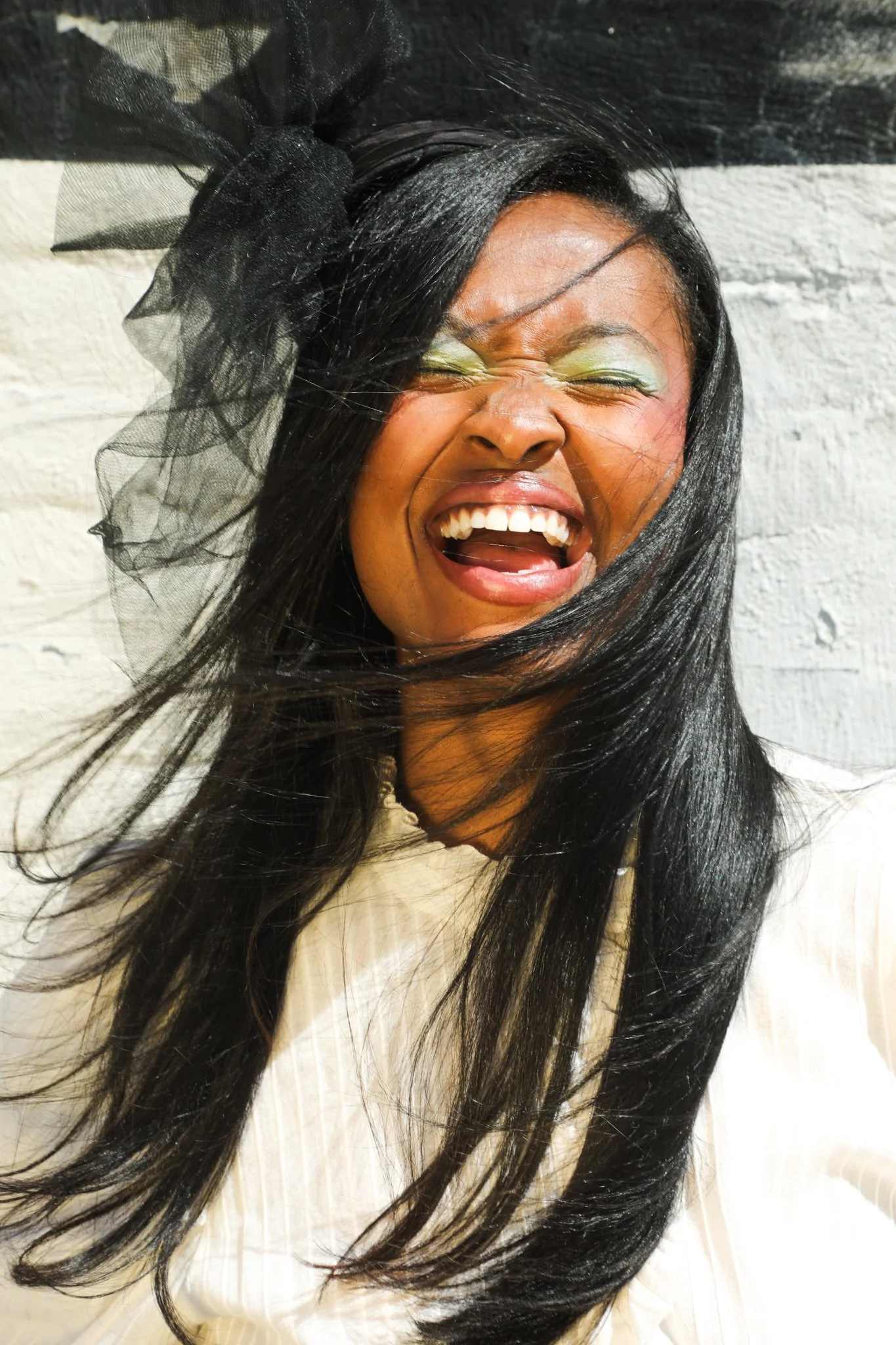 A woman with long black hair, wearing a large black bow with tulle, laughing with closed eyes, in front of a white brick wall.