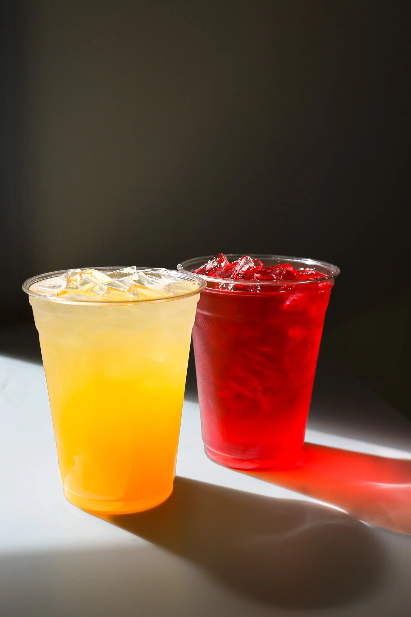 Two beverages in clear plastic cups, one yellow with ice and a lemon slice, one red with ice, on a white surface with shadows and a dark background.