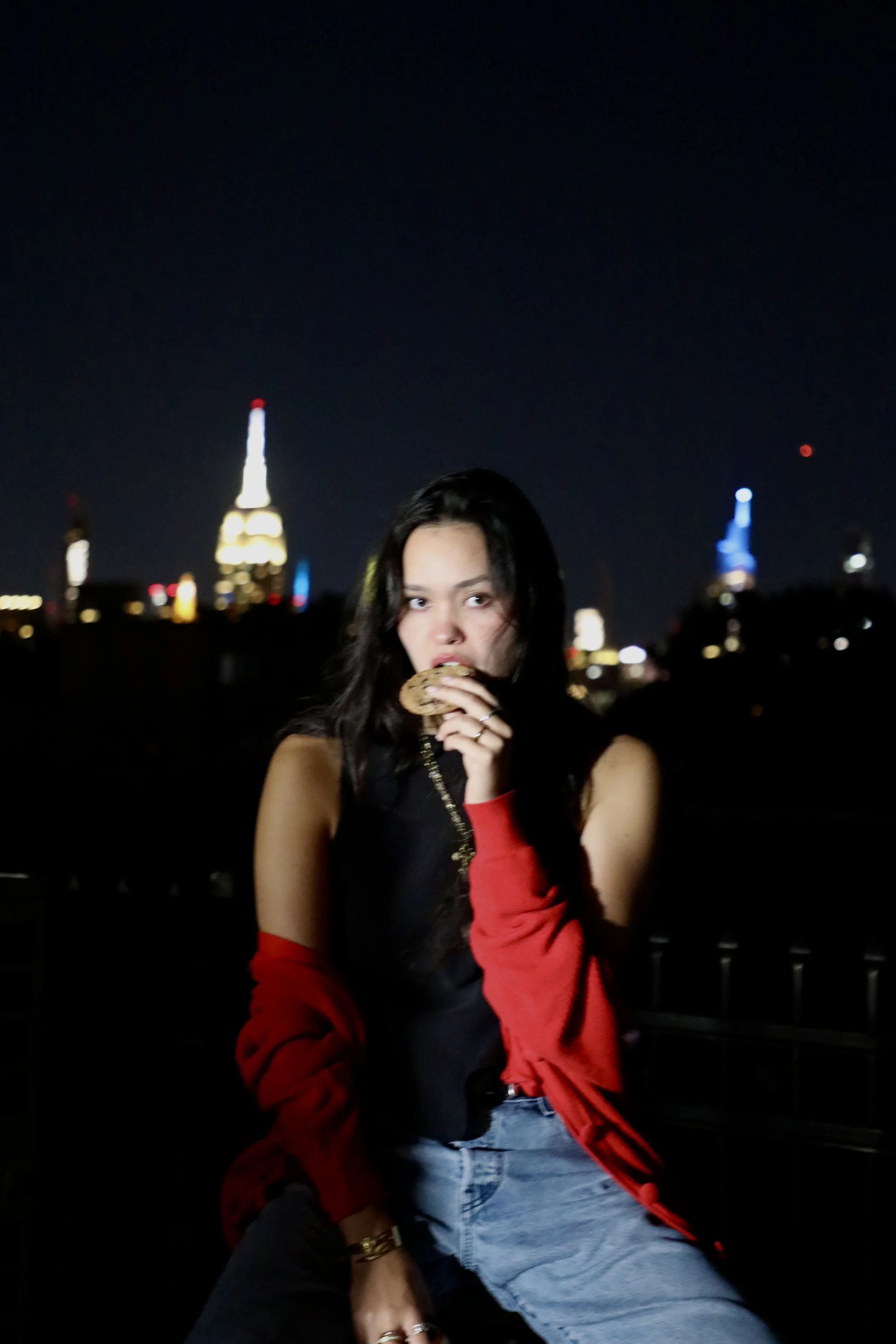 A woman with long dark hair is eating a cookie at night on a rooftop or balcony with a city skyline in the background, including the Empire State Building illuminated.