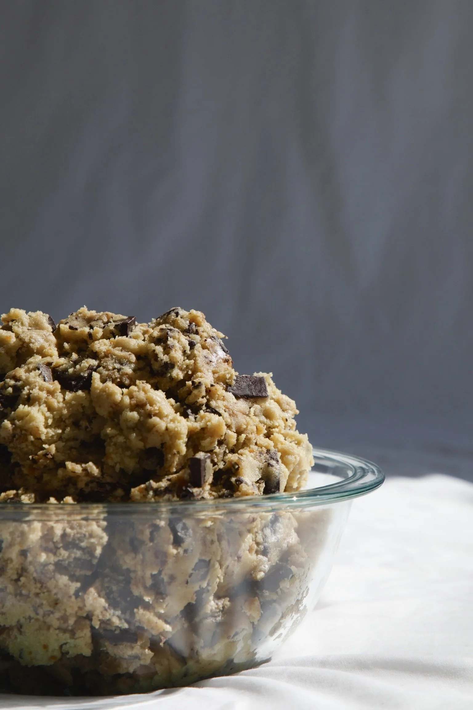 Close-up of chocolate chip cookie dough ice cream served in a clear glass bowl on a white surface with a gray background.