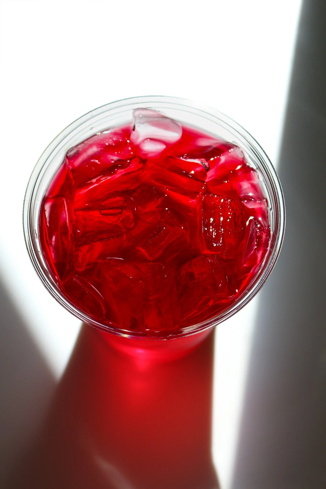 A close-up of a glass filled with red liquid and ice cubes, viewed from above, with a shadow cast on the surface around it.