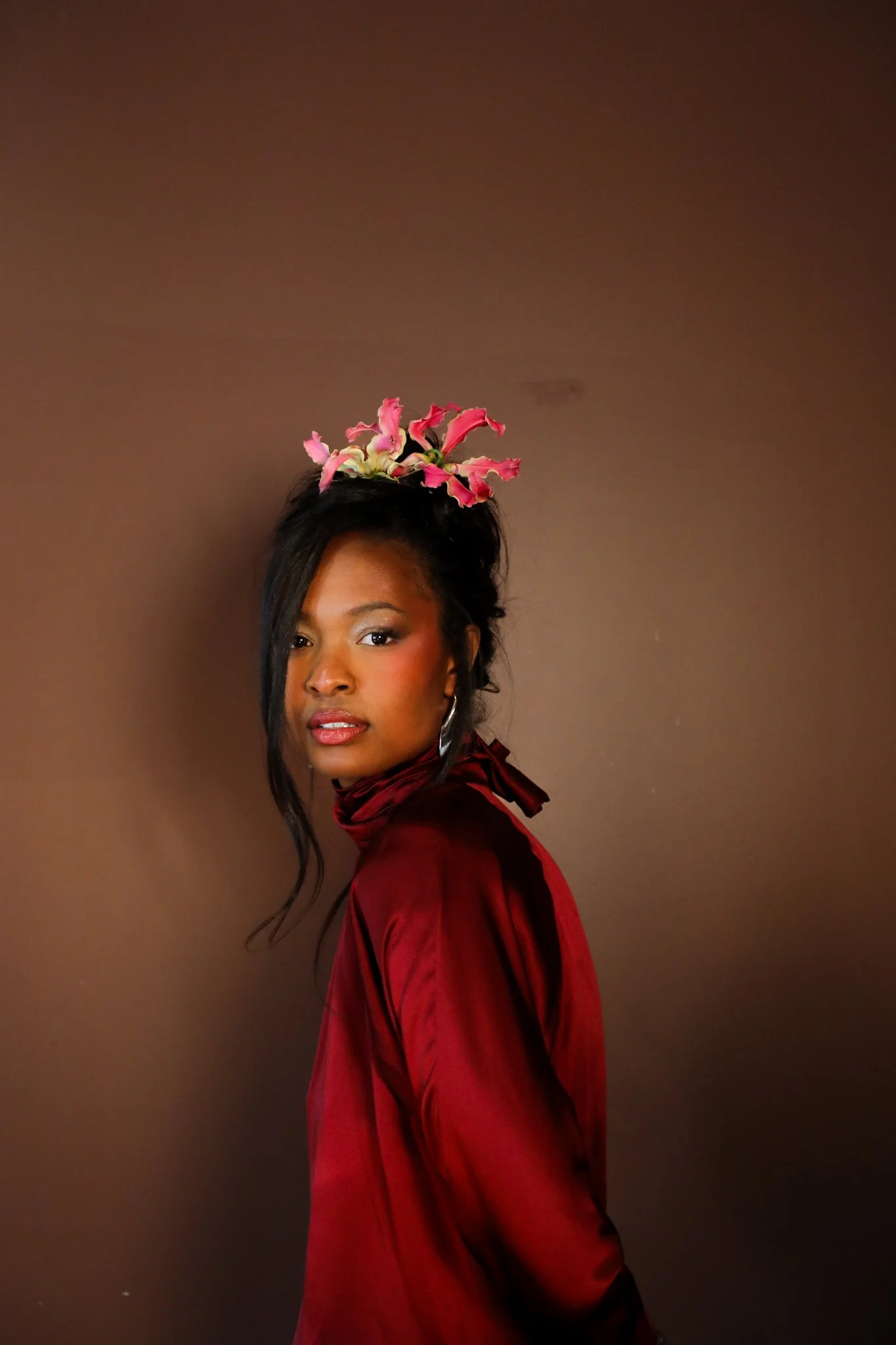 A woman with medium skin tone and dark hair styled in an updo, decorated with pink flowers, wearing a red silk dress, standing against a plain brown background.