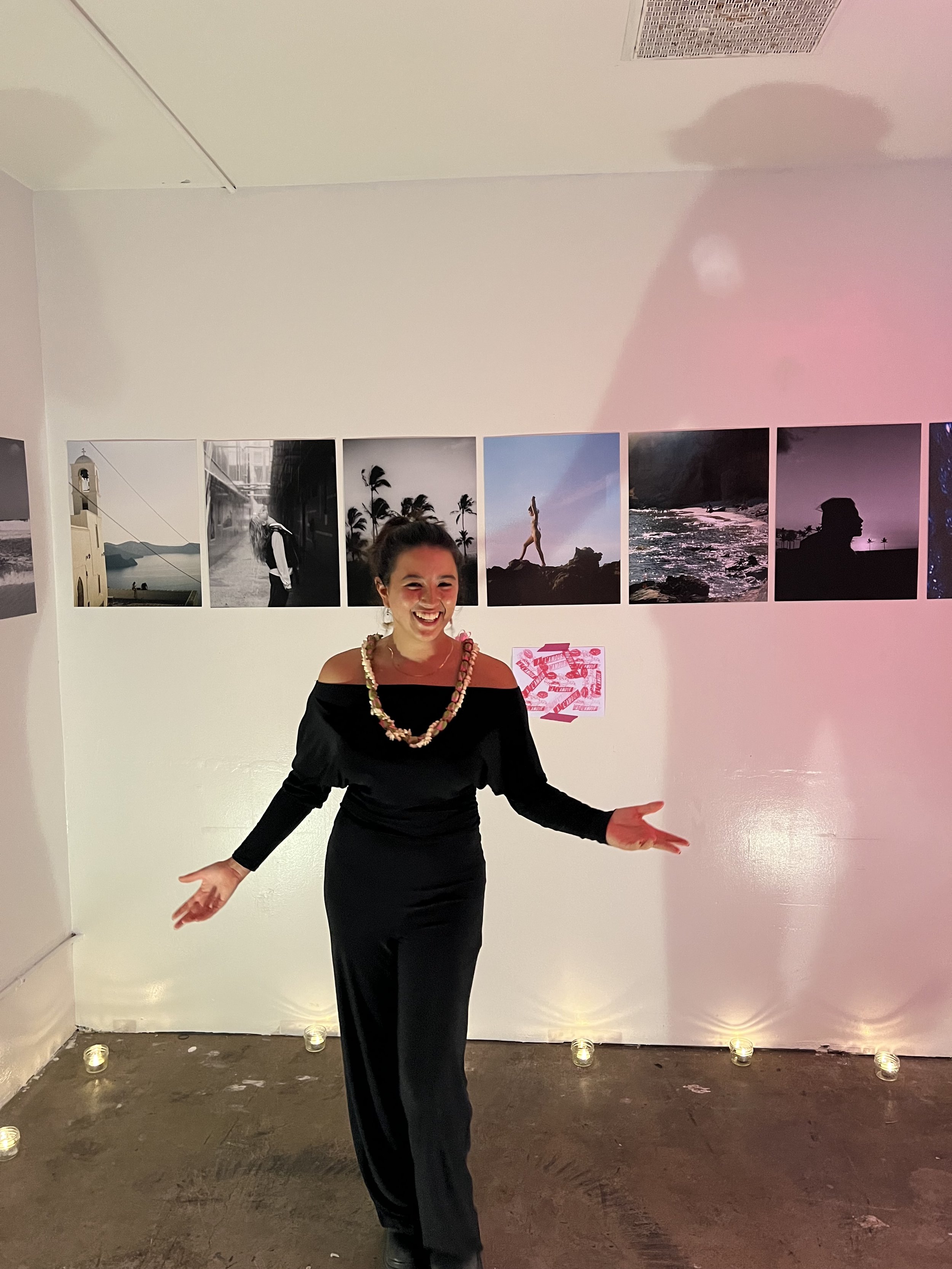A woman standing in front of a wall with framed photographs, smiling and gesturing with her arms out. The photographs on the wall depict various scenes including beach, nature, and urban landscapes. The woman is wearing a black off-shoulder dress and a colorful necklace, and some small candles are lined up at the base of the wall.