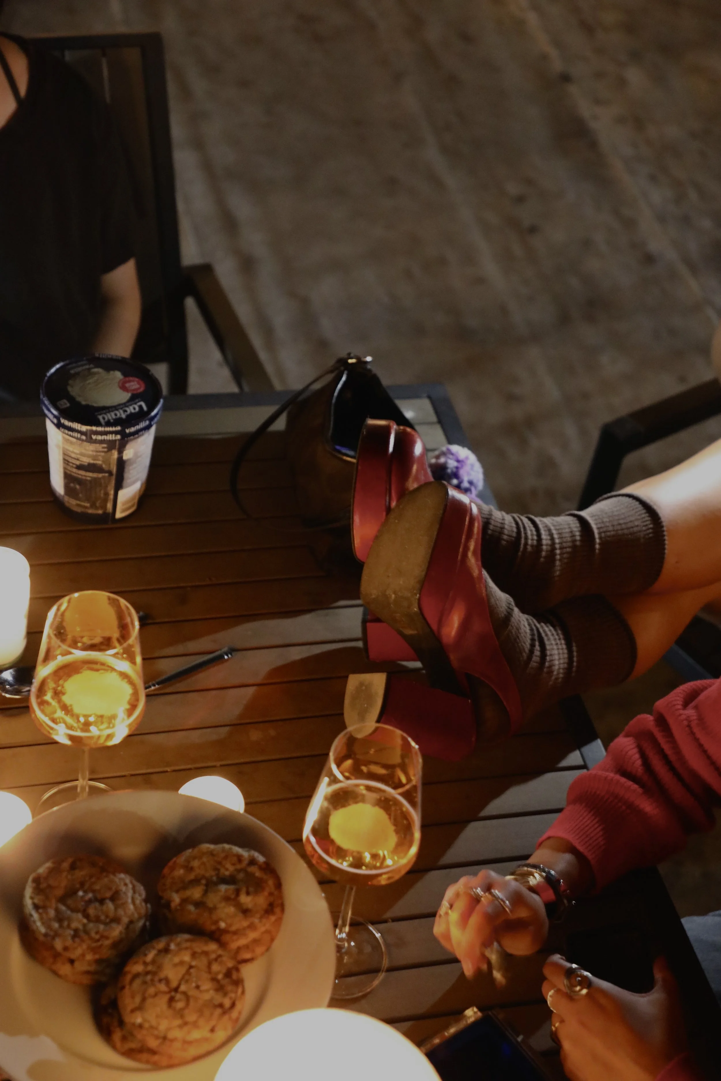 A table with a plate of four cookies, several glasses of a golden-colored drink with lemon slices, and partially visible hands and legs of people wearing red shoes and socks. The setting appears to be indoors with warm lighting.
