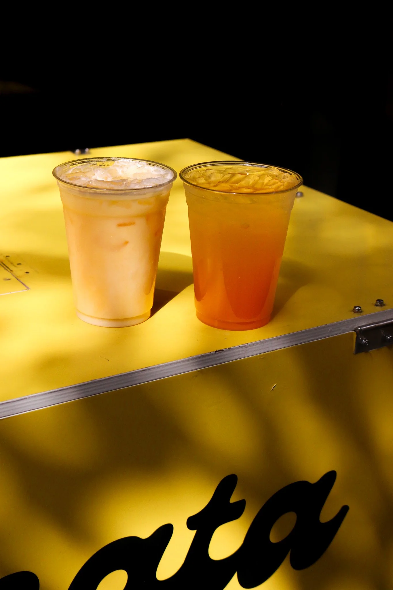 Two cups of iced beverages on a yellow surface with part of black cursive text visible at the bottom.