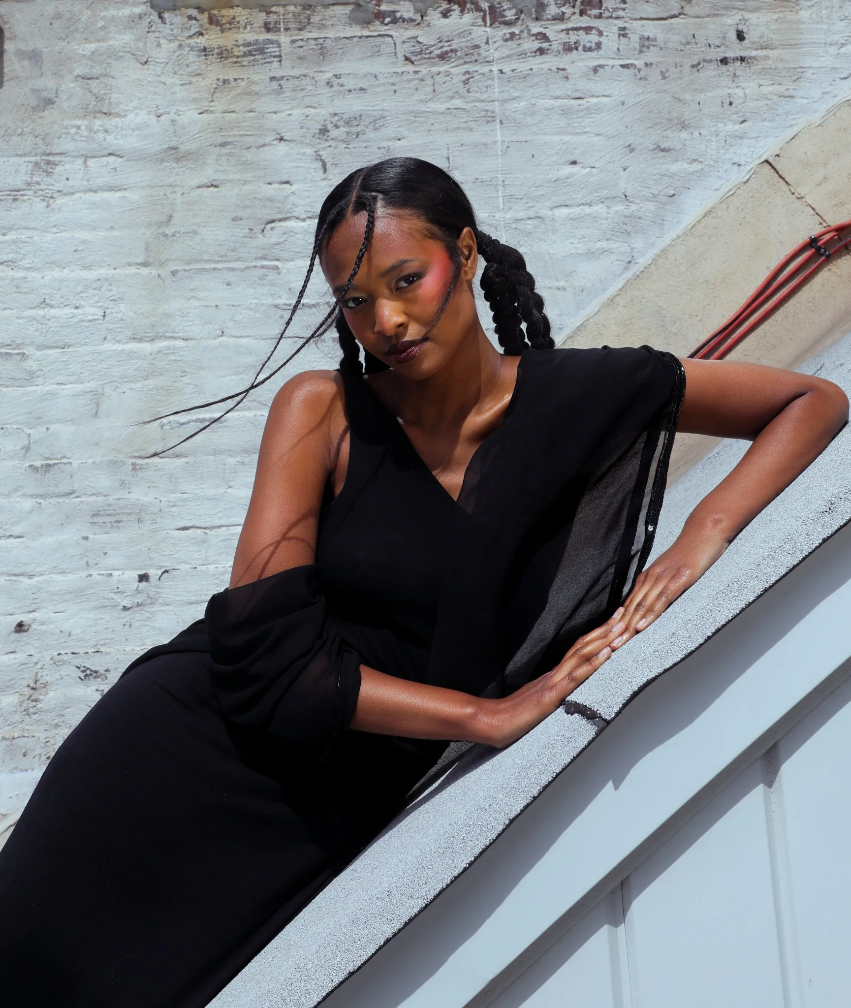 A woman with dark hair styled in two braids, wearing a black top, is leaning against a white brick wall. She has makeup with blush and lipstick, and her expression is confident. The photo is taken at an angle, with part of her arm resting on a ledge and some visible orange cables in the background.