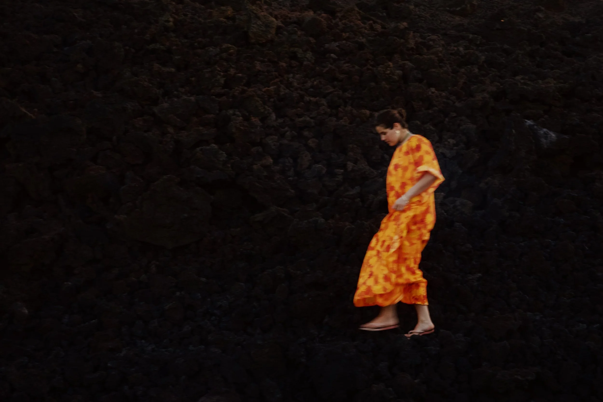 A woman with short dark hair, wearing a bright orange patterned dress or robe, walking barefoot on dark volcanic rocks or terrain.