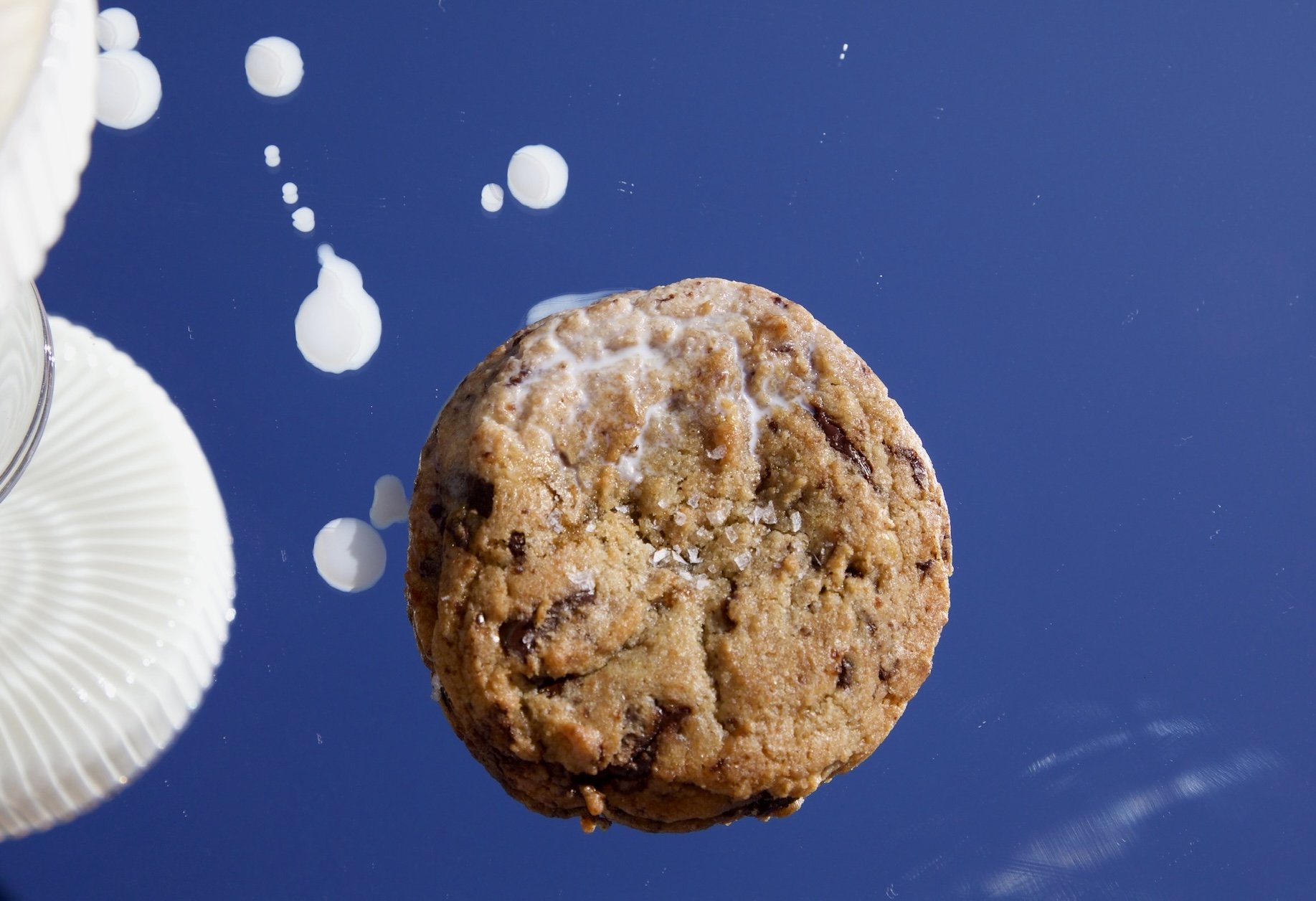 Close-up of a chocolate chip cookie with oats and chocolate chunks on a blue surface, with white round object and edge of a white dish visible.
