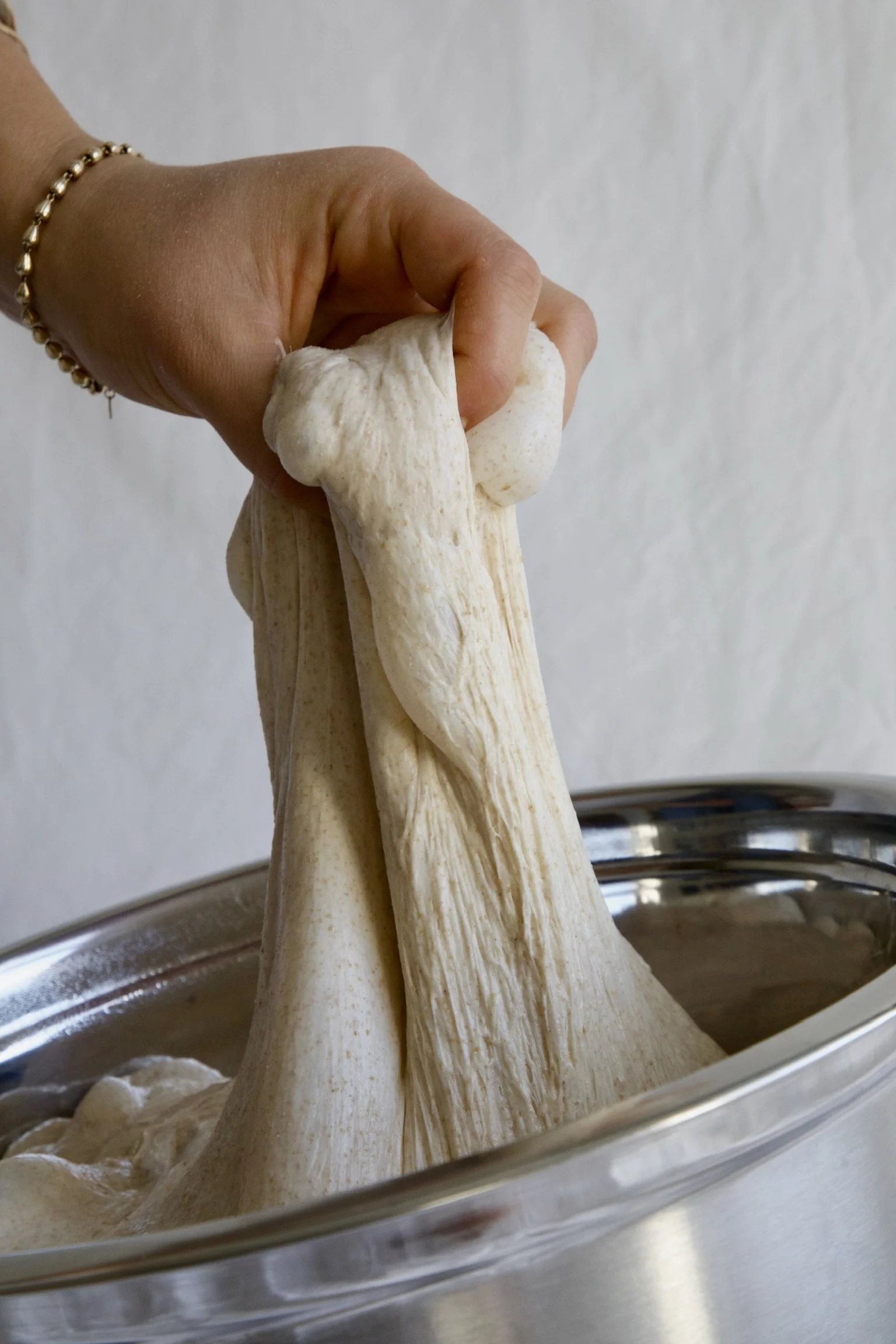 Close-up of a hand kneading dough in a glass bowl, with a plain white wall in the background.