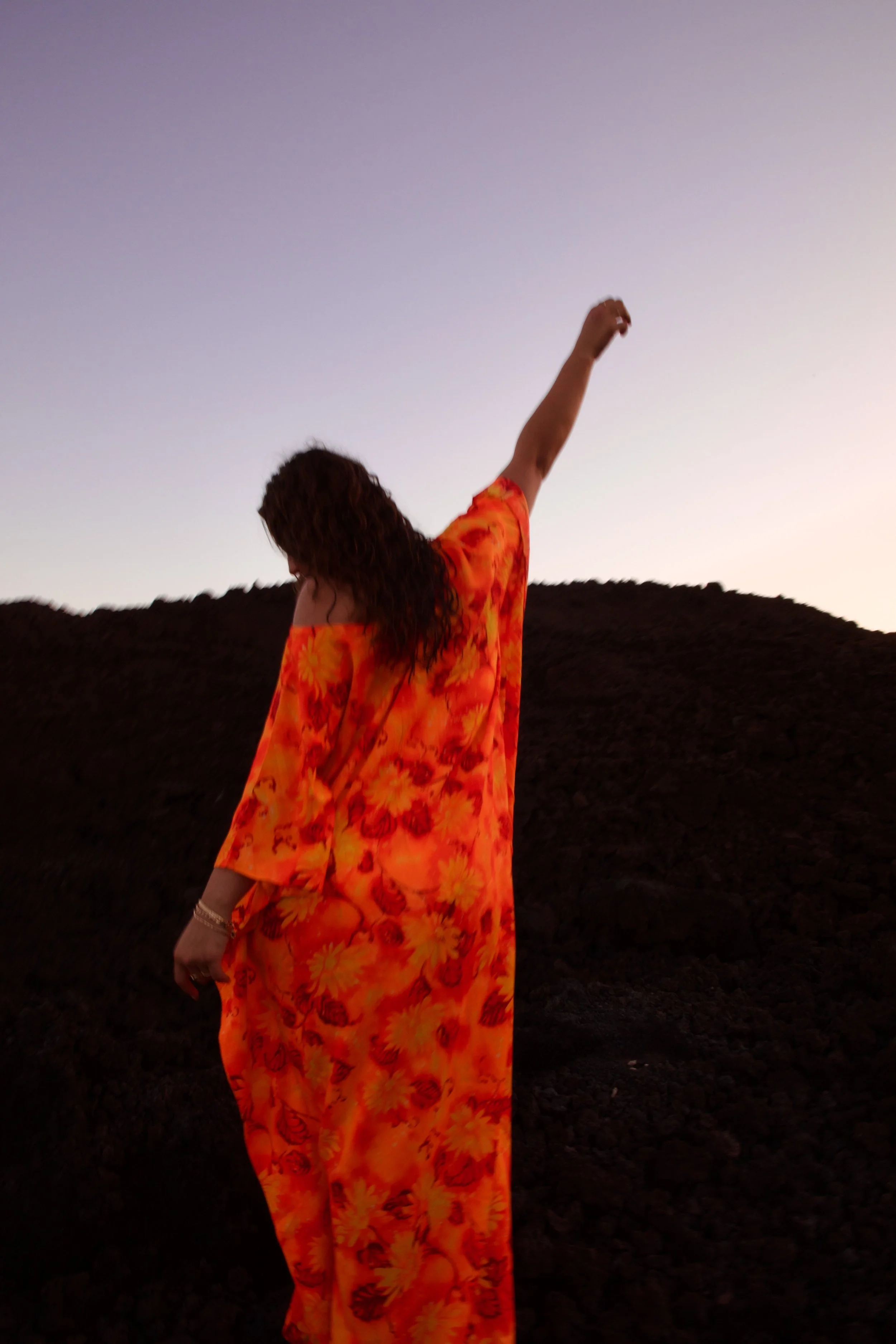 A woman wearing a bright orange and red floral dress, standing outdoors at sunset, with one arm raised and a mountain range in the background.