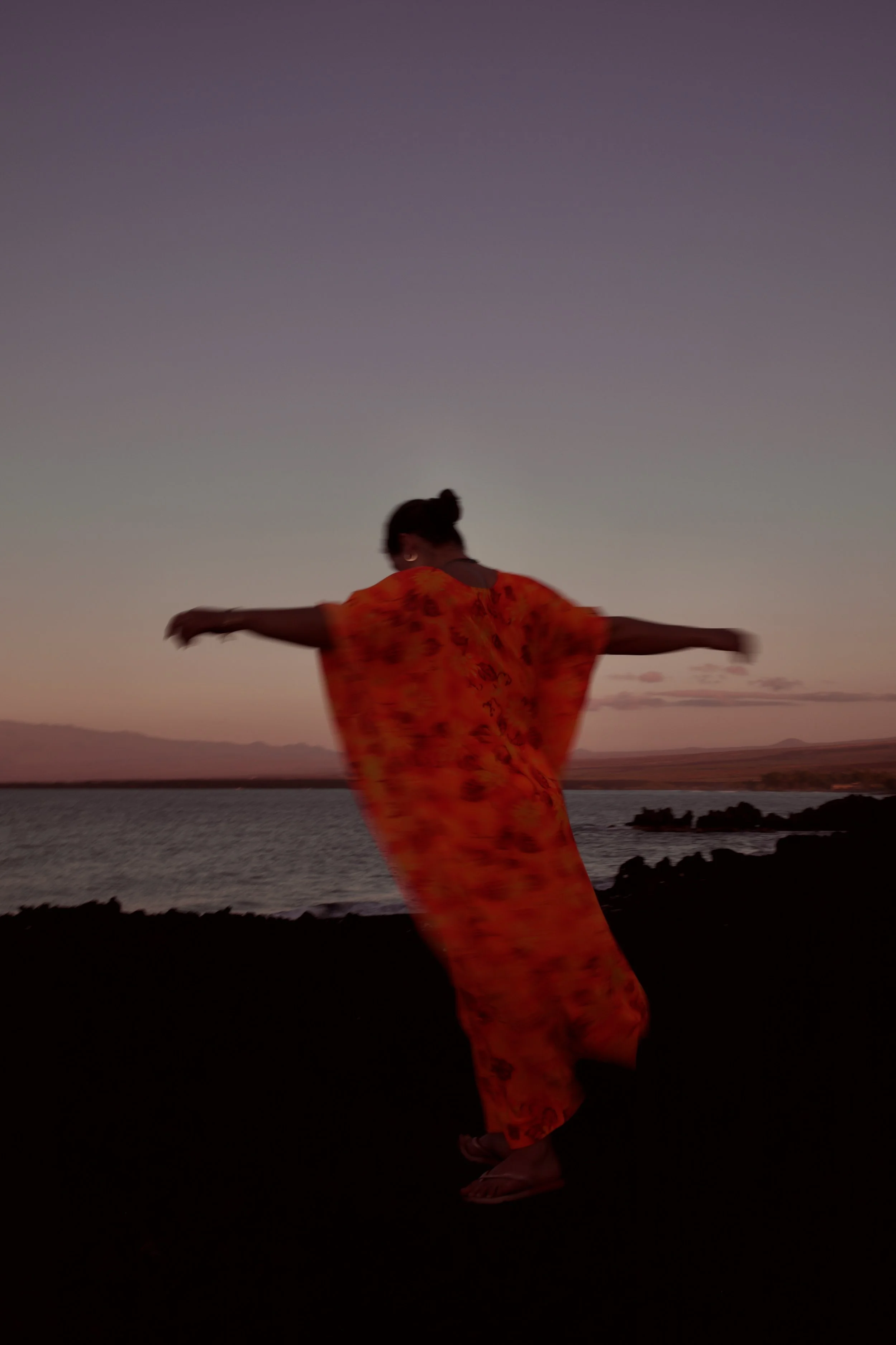 A woman in a flowing orange dress with floral patterns standing by a body of water at sunset with her arms outstretched.
