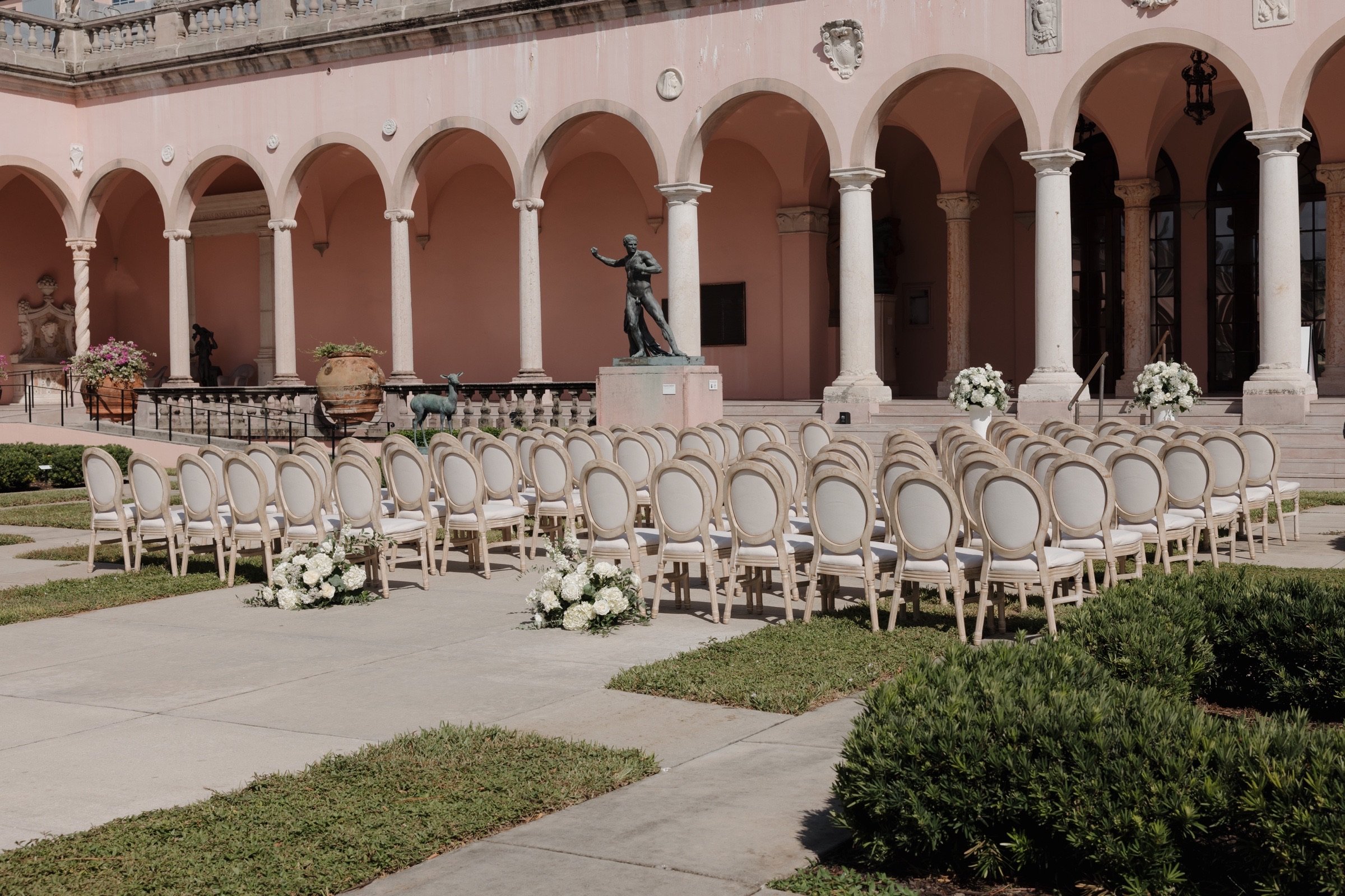 Sunlit pink courtyard at The Ringling in Sarasota, with pale arches, rows of light chairs, and white floral arrangements.