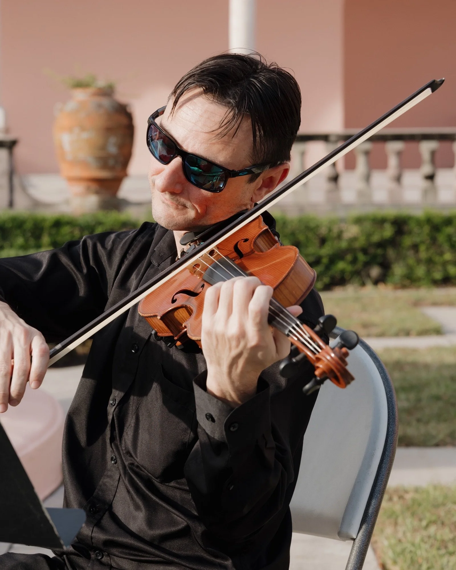 Violinist Vasi seated and playing violin in a sunlit pink courtyard at The Ringling in Sarasota.