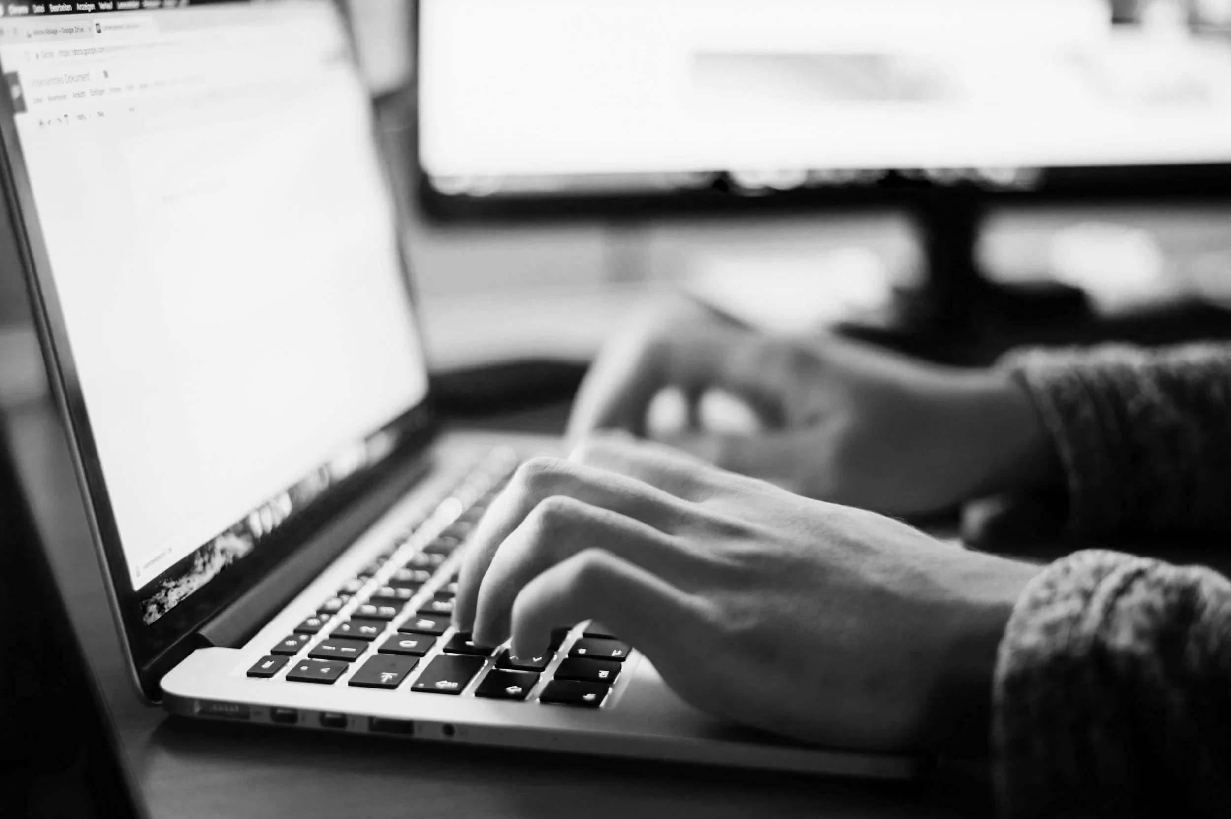 Close-up of hands typing on a laptop keyboard with a blurred computer screen in the background in black and white.