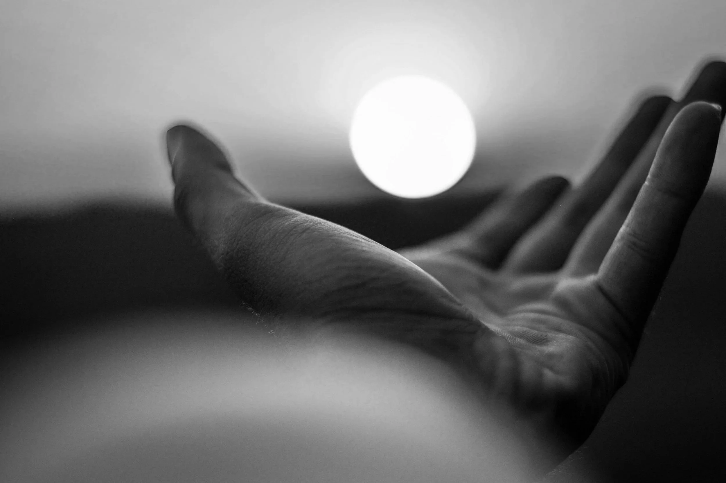 Black and white photo of a person's hand reaching towards a bright, glowing circular light source in the background.