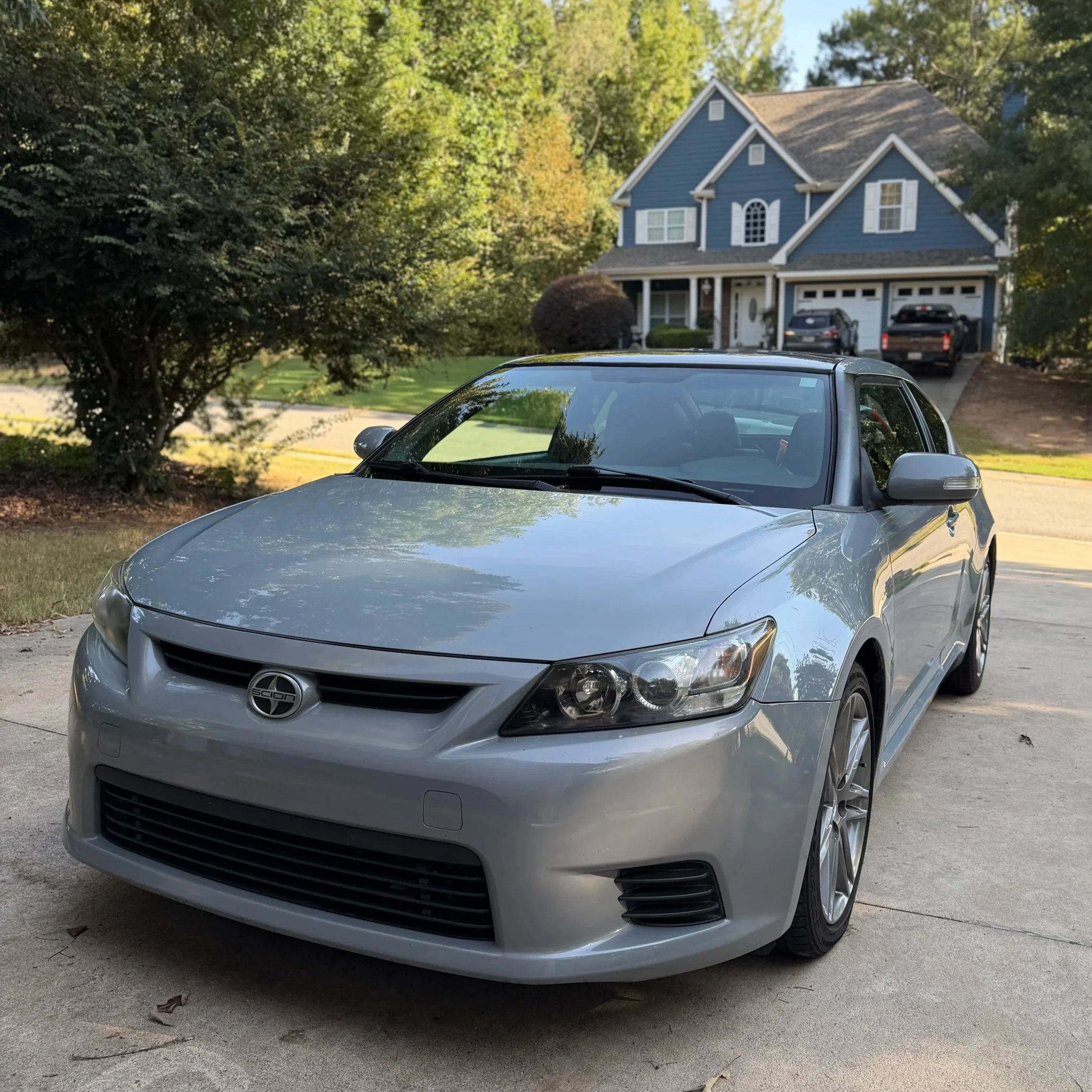 Front view of a silver sports car parked on a driveway in front of a blue two-story house with white trim and a garage, surrounded by trees.