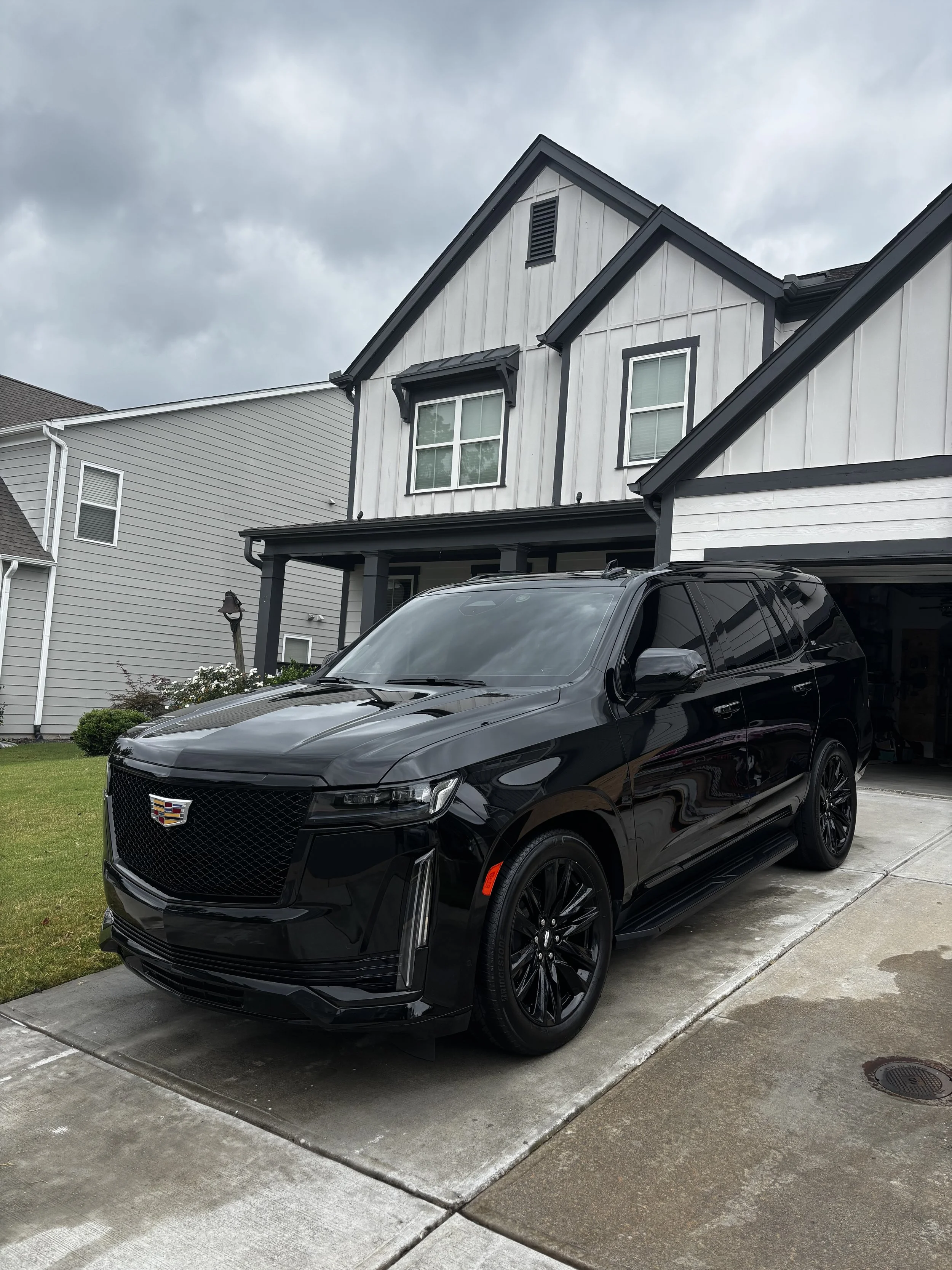 Black Cadillac SUV parked in driveway in front of a modern house with gray and white exterior, cloudy sky overhead.