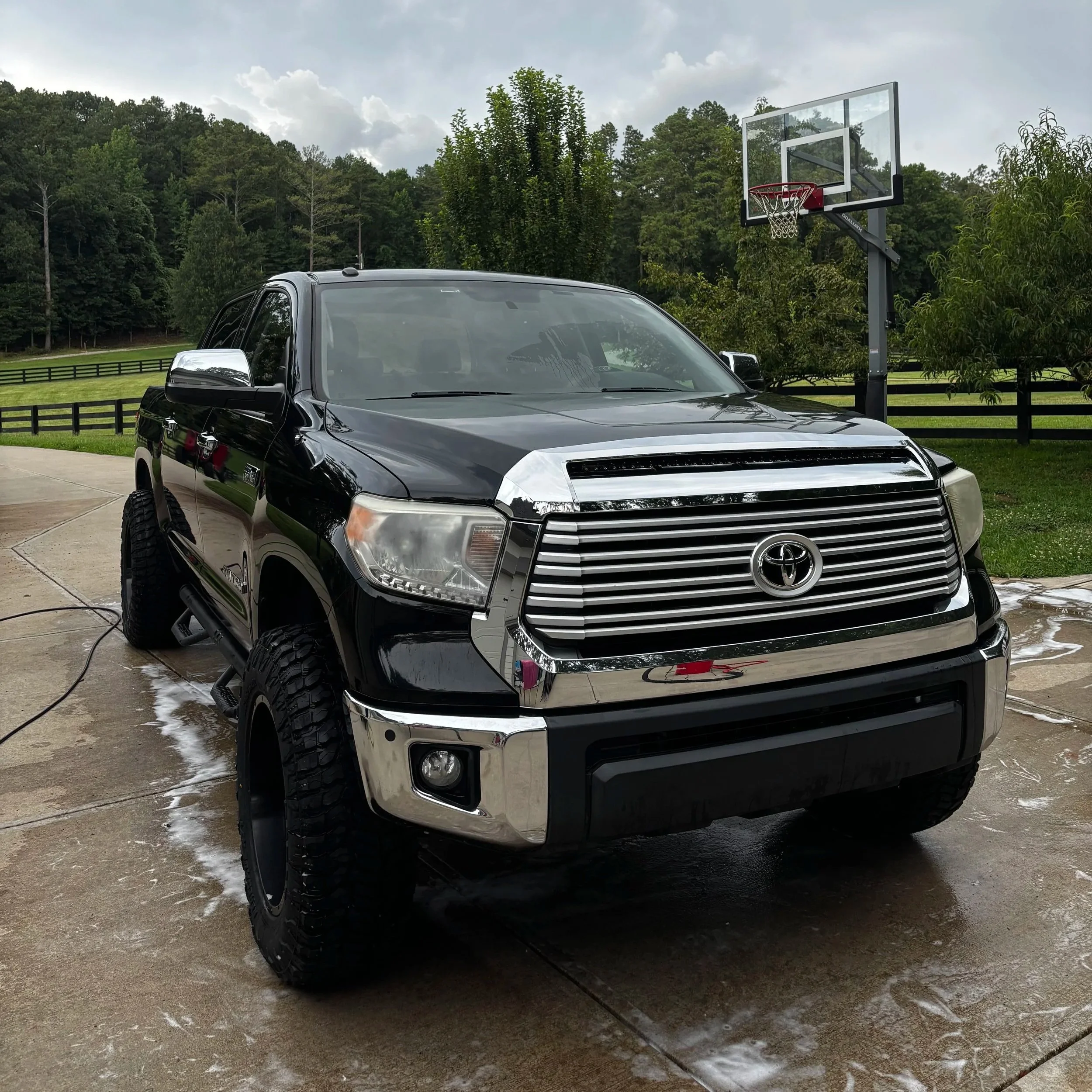A black Toyota Tundra pickup truck parked on a driveway with a sport basketball hoop nearby and a background of green trees and a cloudy sky.
