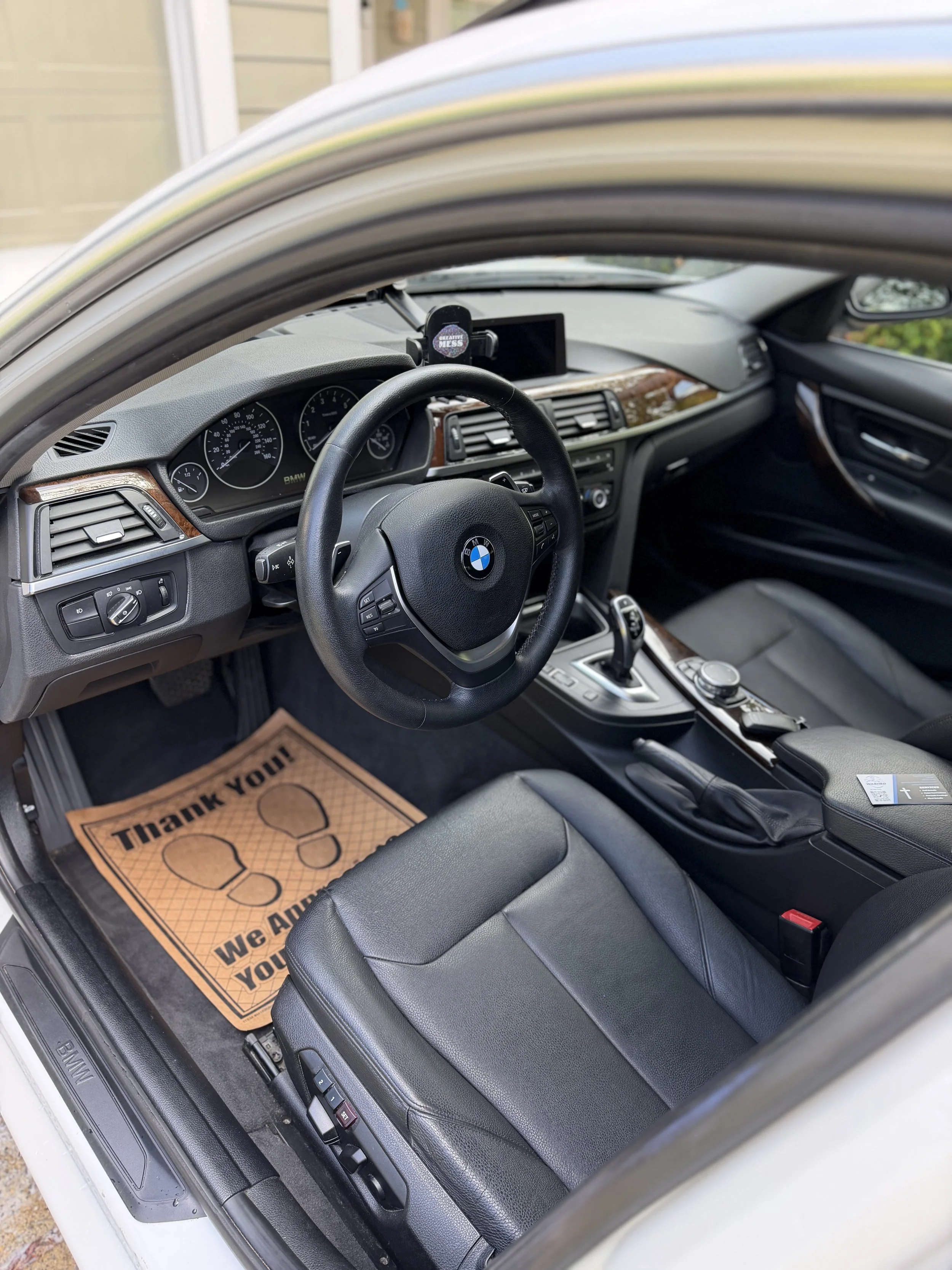 Interior of a BMW car showing the steering wheel, dashboard, center console, and black leather seats, with a paper floor mat that reads 'Thank You' and 'We Appreciate Your Business'.
