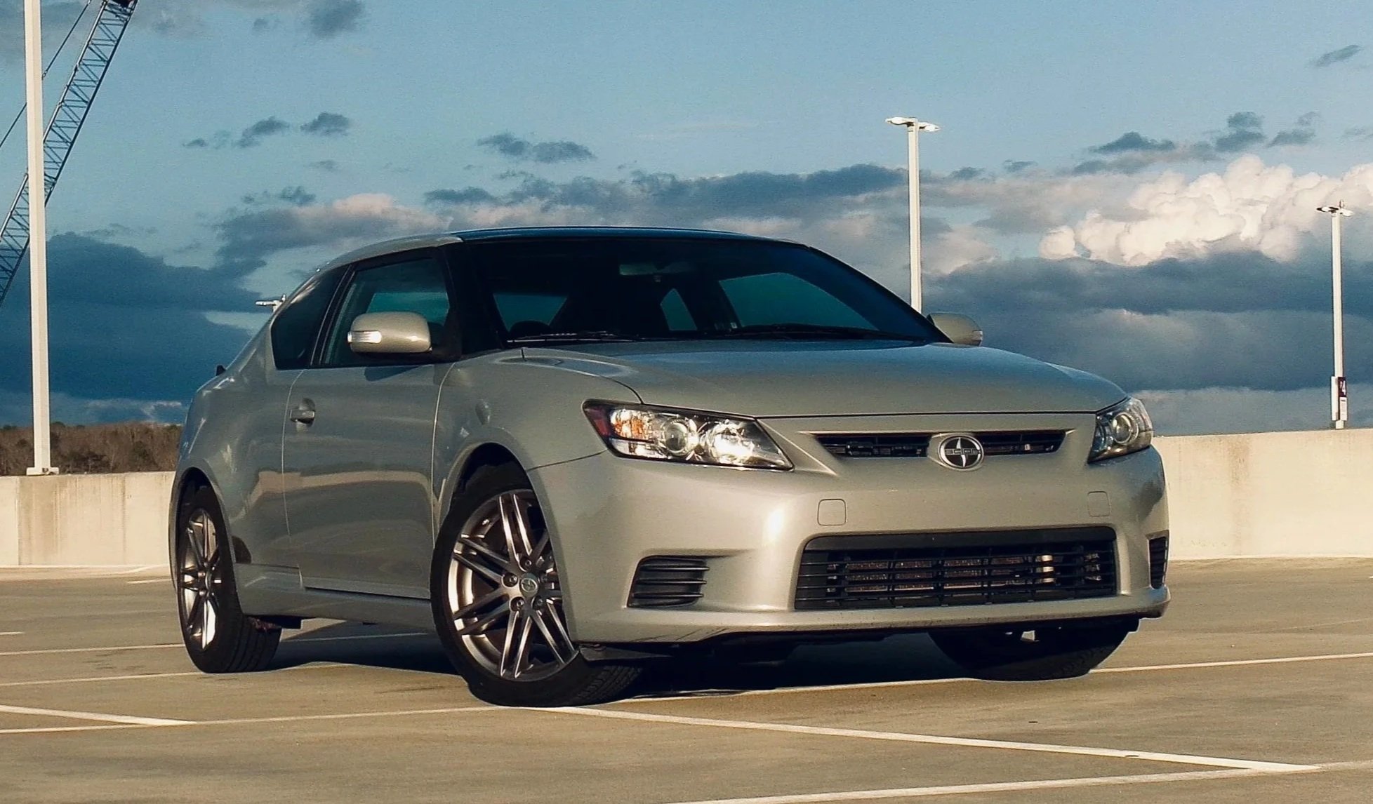 A silver Scion tC car parked in an empty outdoor parking lot under a partly cloudy sky.