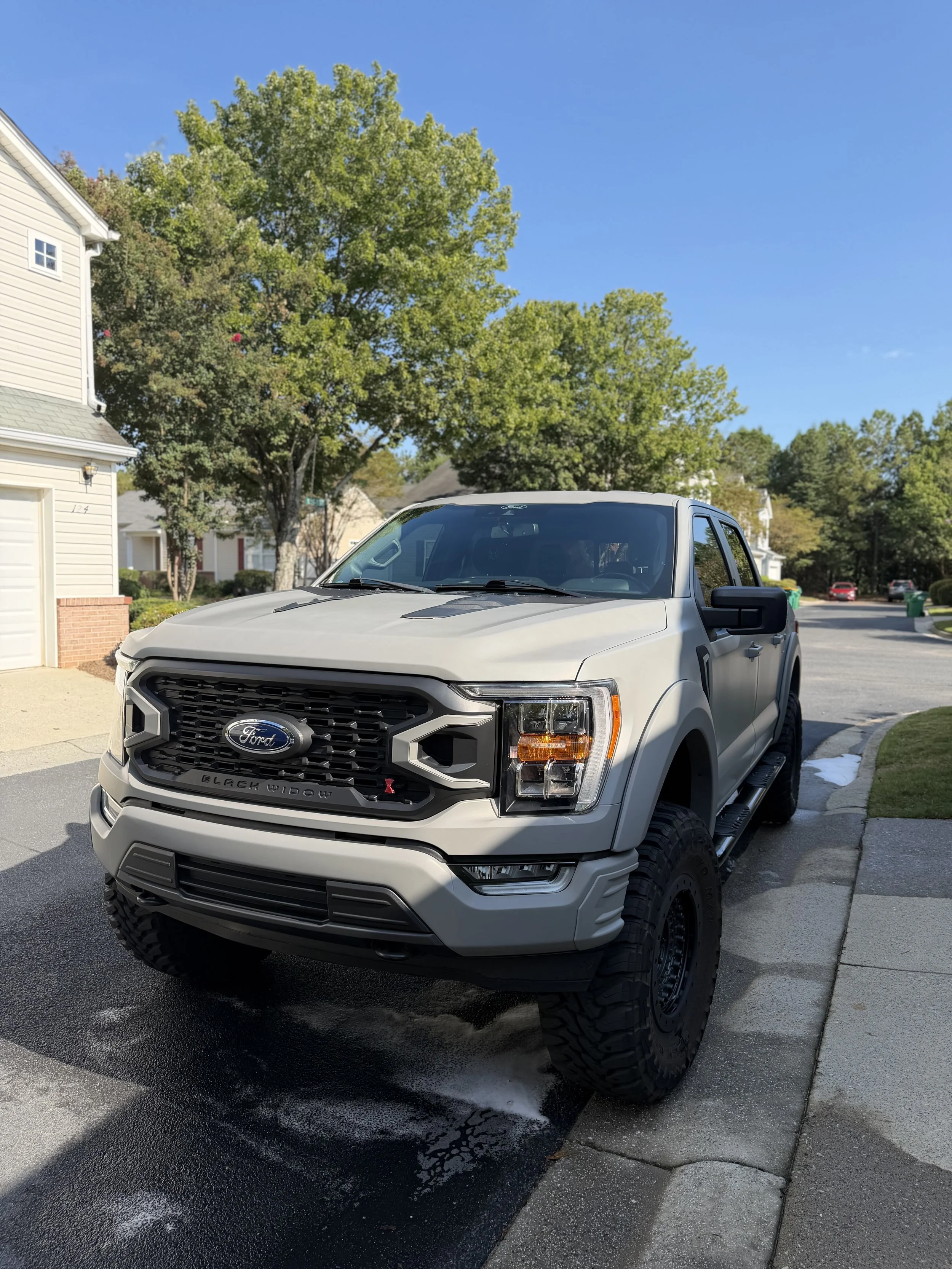 A white Ford pickup truck parked on a residential street with a large tree, houses, and a clear blue sky in the background.