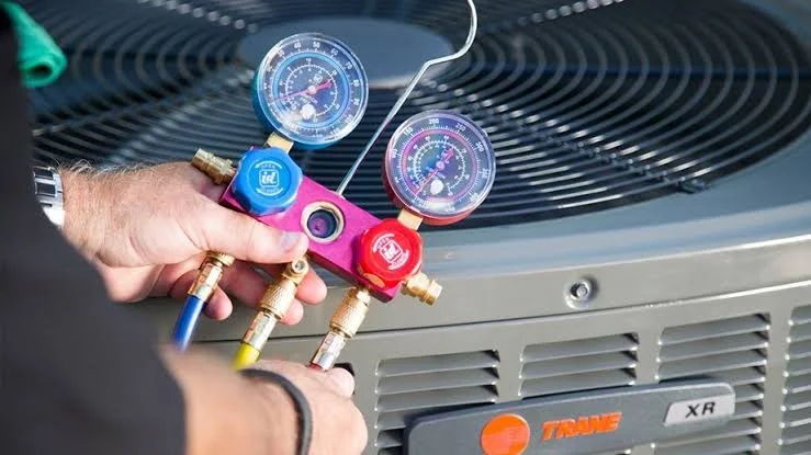 A technician using a manifold gauge set with blue and red gauges to diagnose an air conditioning unit.