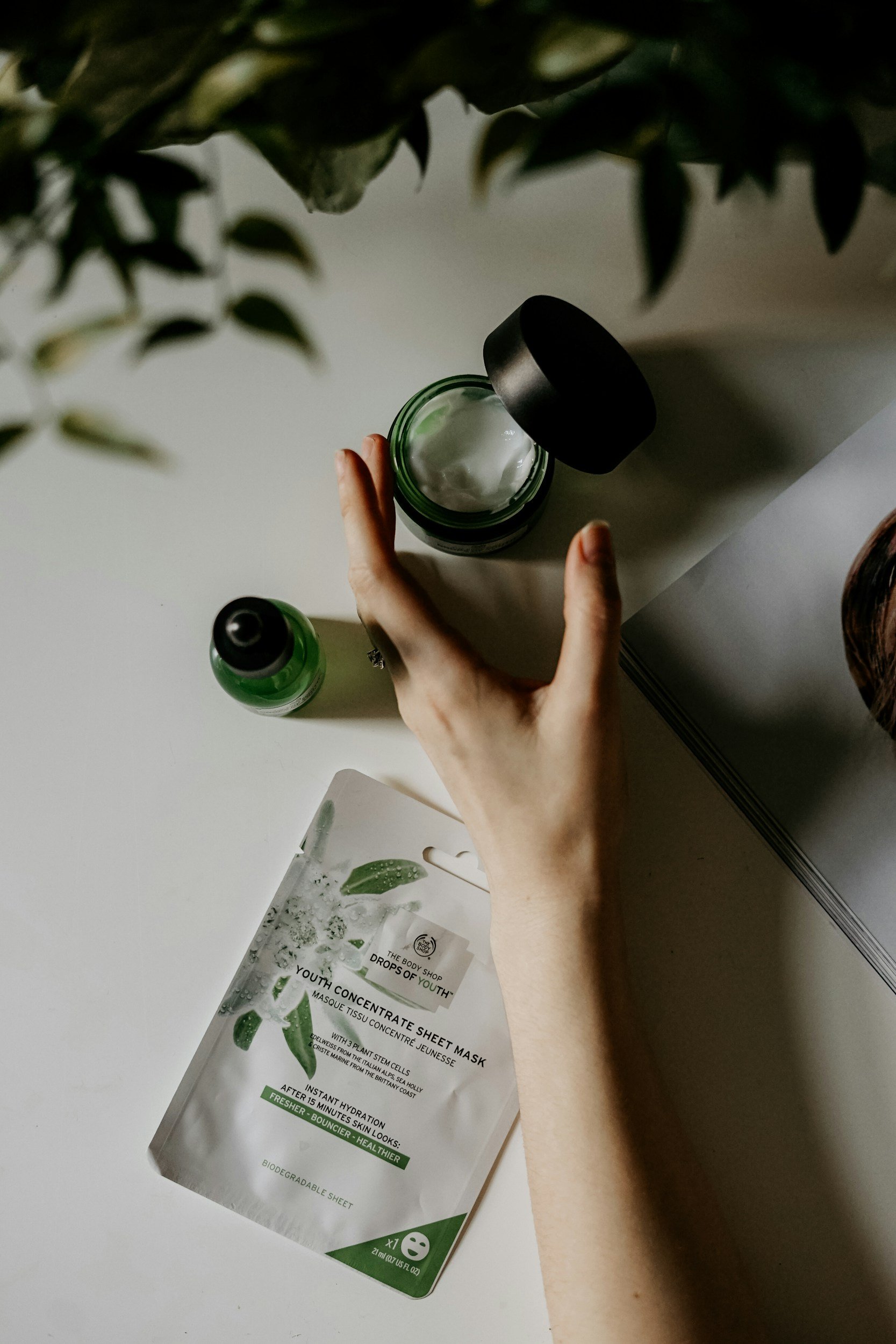 A hand holding a jar of green cream or moisturizer on a white surface. Green plant leaves hanging over the frame. A packet labeled 'The Body Shop Drops of Youth' sheet mask on the table.