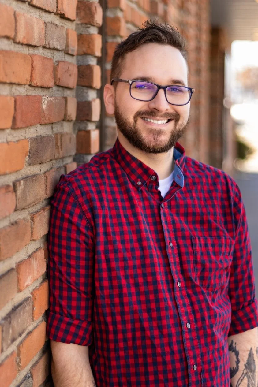 A smiling man with glasses and a beard, wearing a red and blue checkered shirt, standing next to a brick wall outdoors.