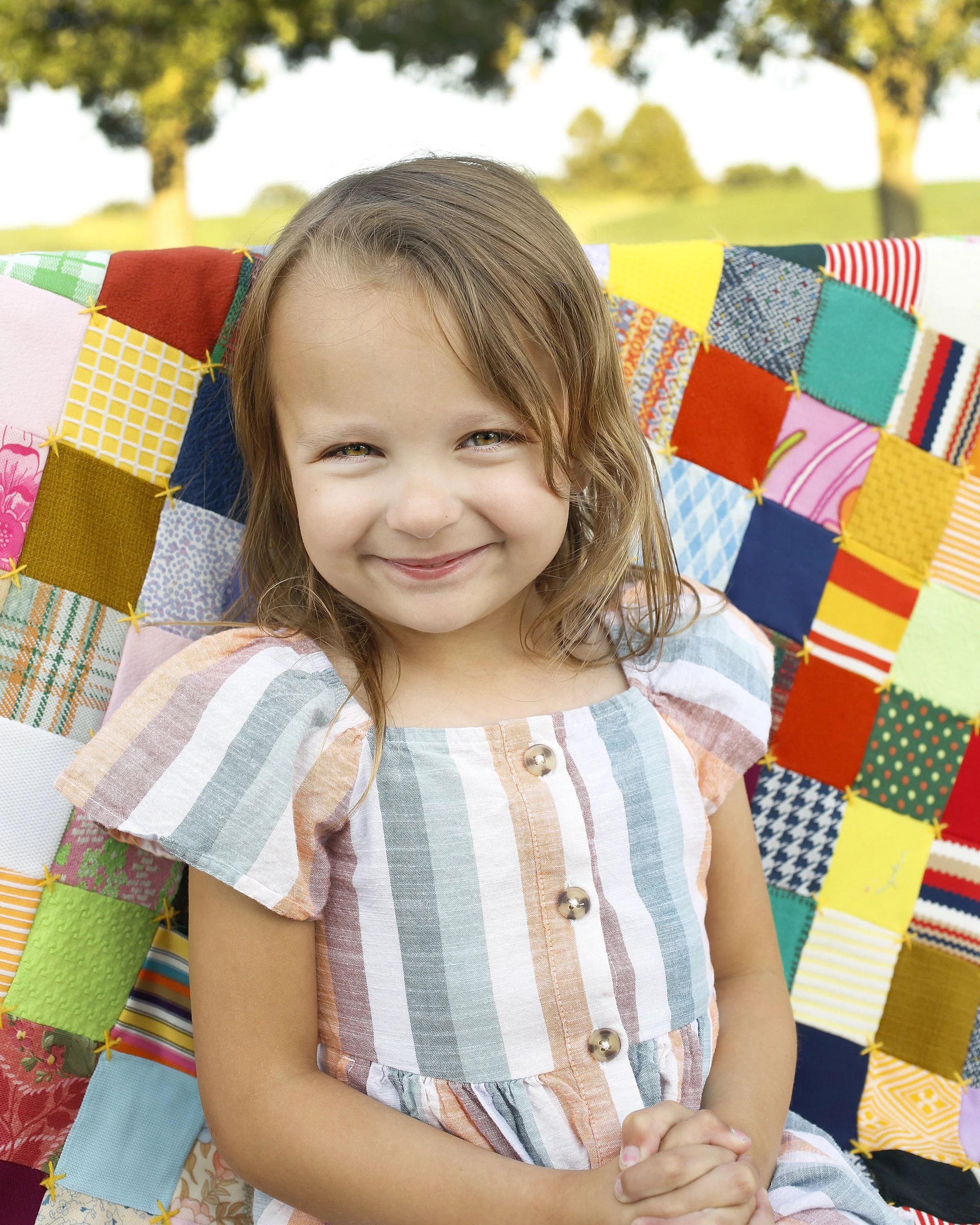 A young girl with light brown hair and a smile, sitting outdoors with a colorful fabric quilt behind her. The quilt features various bright and patterned squares, and there's a background of green trees and a sunny sky.