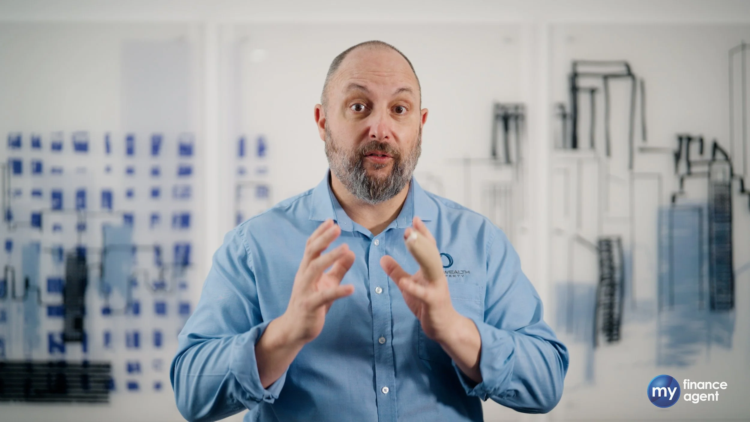 A man with a beard and short hair, wearing a light blue collared shirt, gesturing with his hands while speaking in a bright office or commercial space with white walls, blue and black display frames, and equipment in the background.